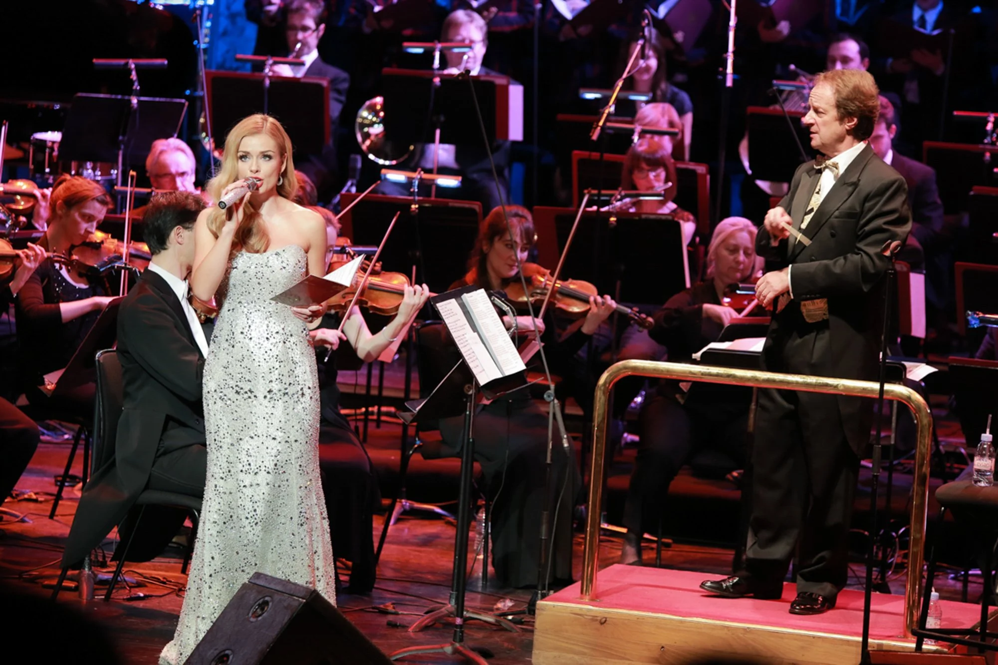 A woman in a white, sparkly, strapless gown singing into a microphone, with a man in a tuxedo sitting beside her, during a performance with an orchestra behind them on stage.