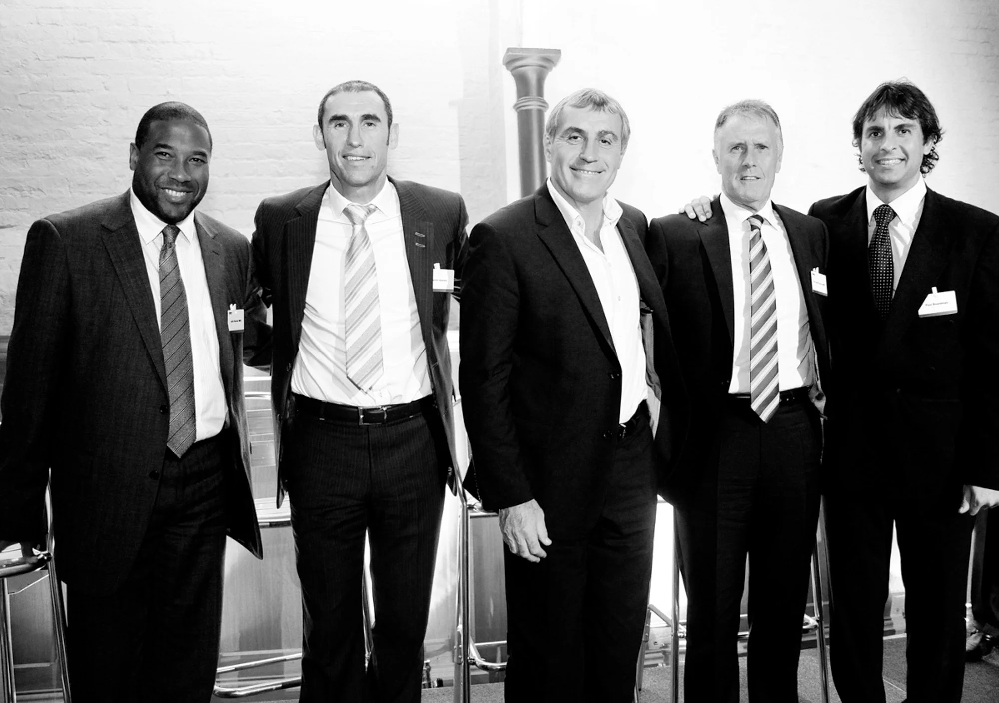 Six men in formal business attire standing together indoors, facing the camera, with a brick wall background.