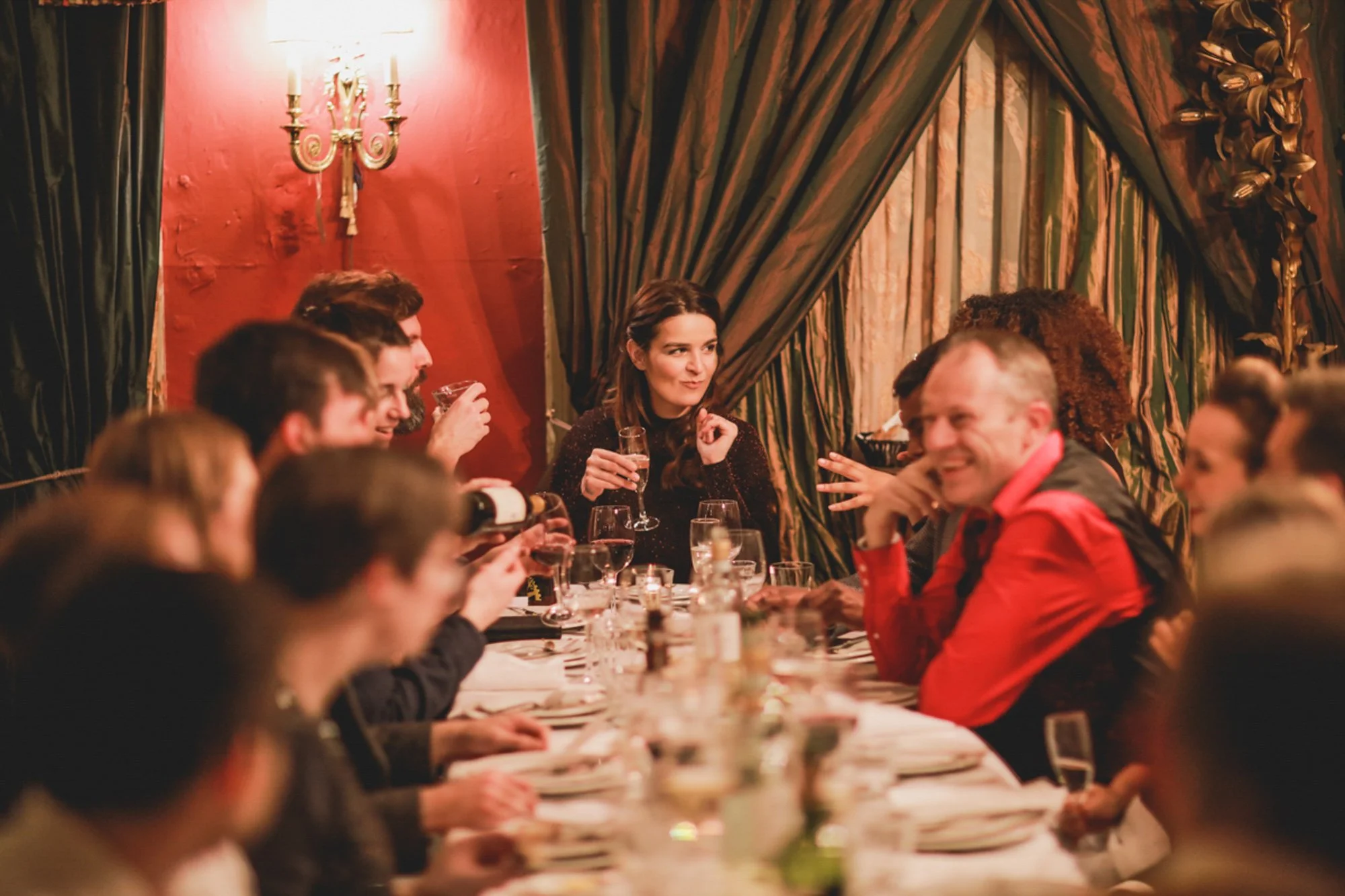 People gathered around a dinner table with drinks, in a warmly lit, ornate room with curtains and red walls, celebrating or socializing.