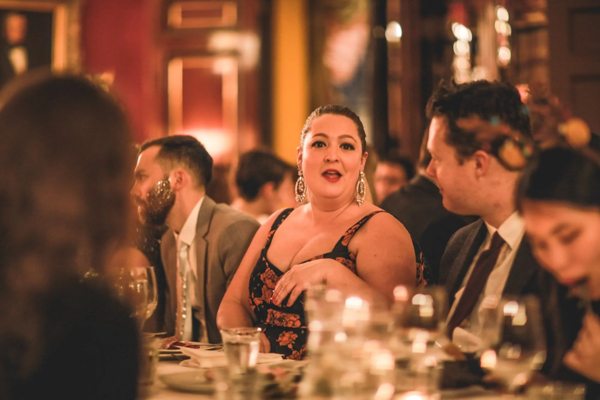 A woman in a floral dress and large earrings speaking to a man in a suit at a dinner event.
