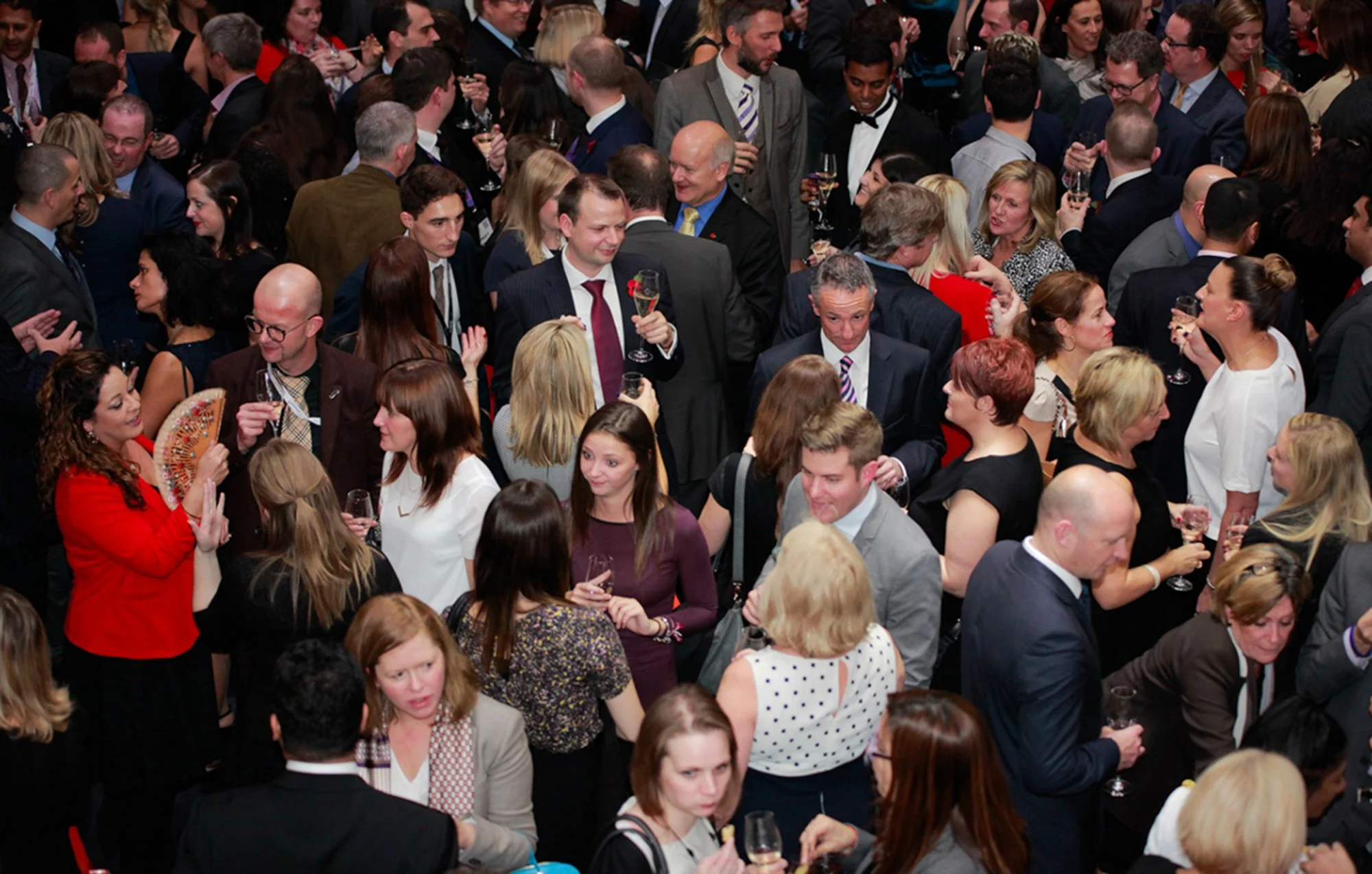 A large crowd of people dressed in formal attire gathered at a social event, holding glasses of champagne and engaging in conversations.