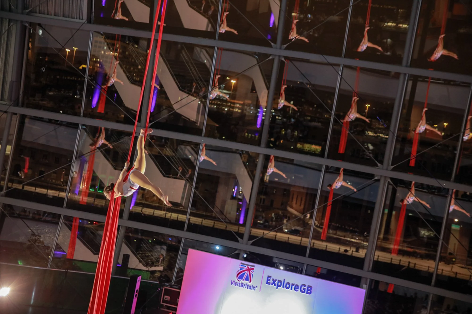 Aerial silk performers demonstrating aerial silks in front of a glass building with multiple reflections at night. An LED screen displaying 'Visit Britain' and 'ExploreGB' is visible below.