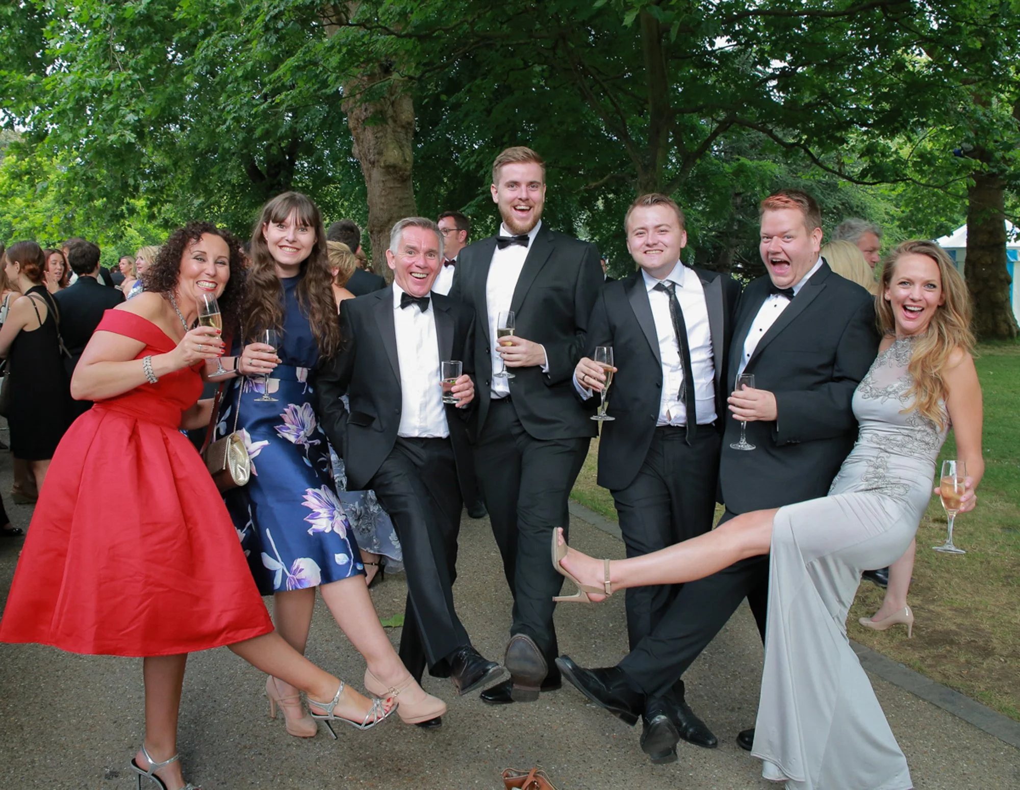 A group of seven people dressed in formal attire, smiling and holding champagne glasses at an outdoor event, with green trees and other guests in the background.