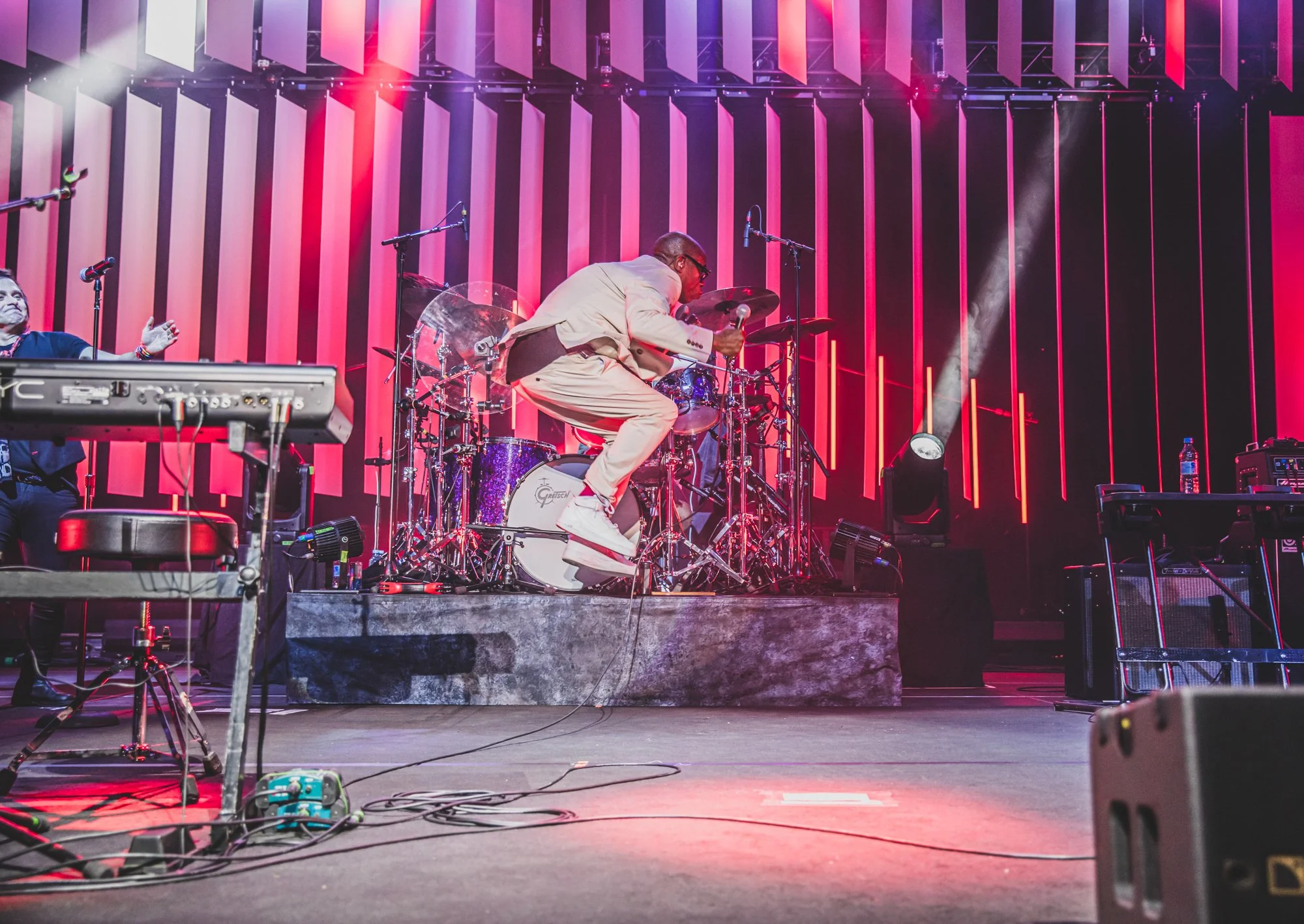 Musician jumping on stage during a concert with a brightly lit colorful background and musical equipment.