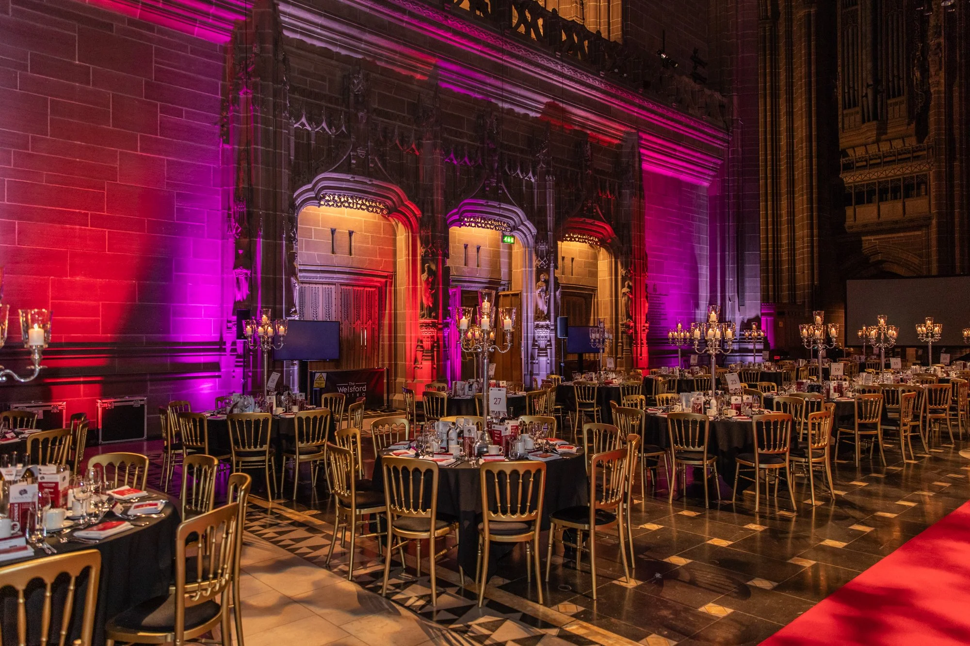 Elegantly decorated event hall with round tables covered in black tablecloths, surrounded by gold chairs, and tall candelabras with lit candles. The grand stone walls are lit with colorful pink and purple lights, highlighting intricate architectural