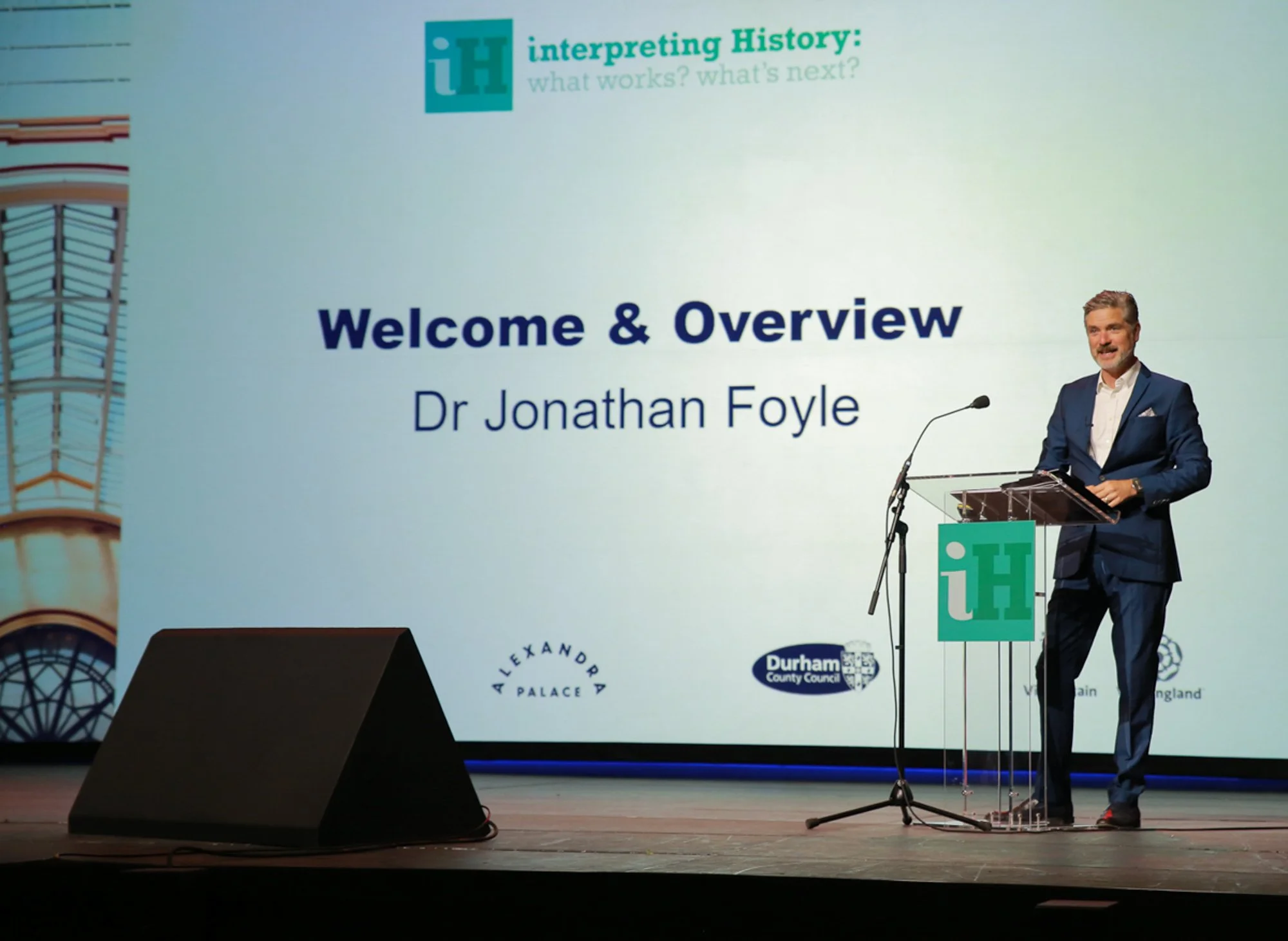 A man in a dark blue suit standing at a clear podium with a microphone, giving a presentation on stage. Behind him is a large screen displaying a slide titled 'Welcome & Overview' with the name 'Dr Jonathan Foyle,' logos for Alexandra Palace, Durham