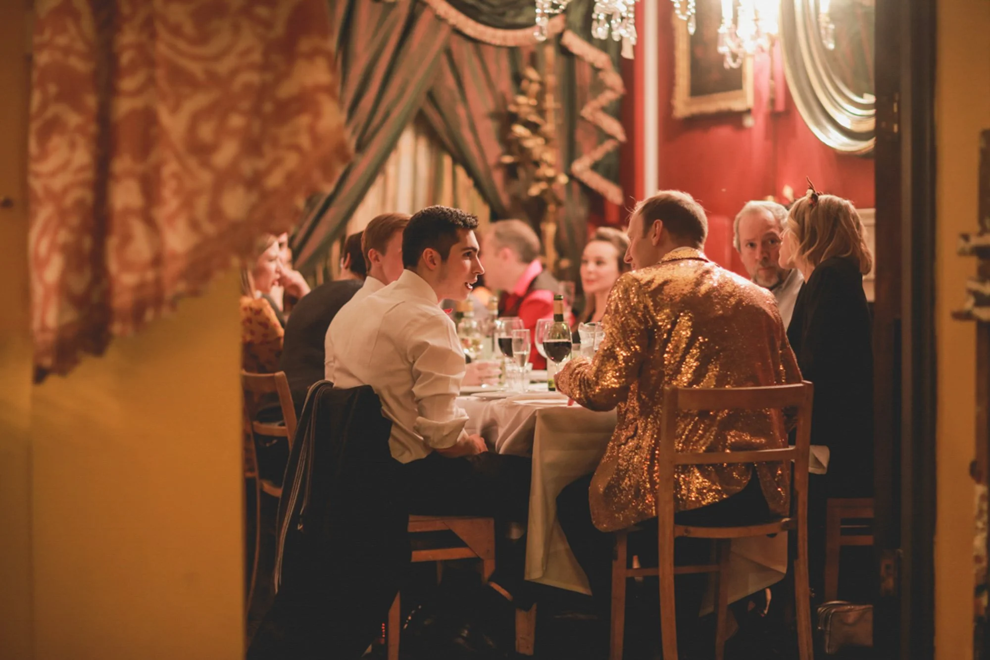 People gathered around a dining table in a warmly decorated restaurant, engaged in conversation and enjoying drinks, with ornate curtains and a chandelier in the background.