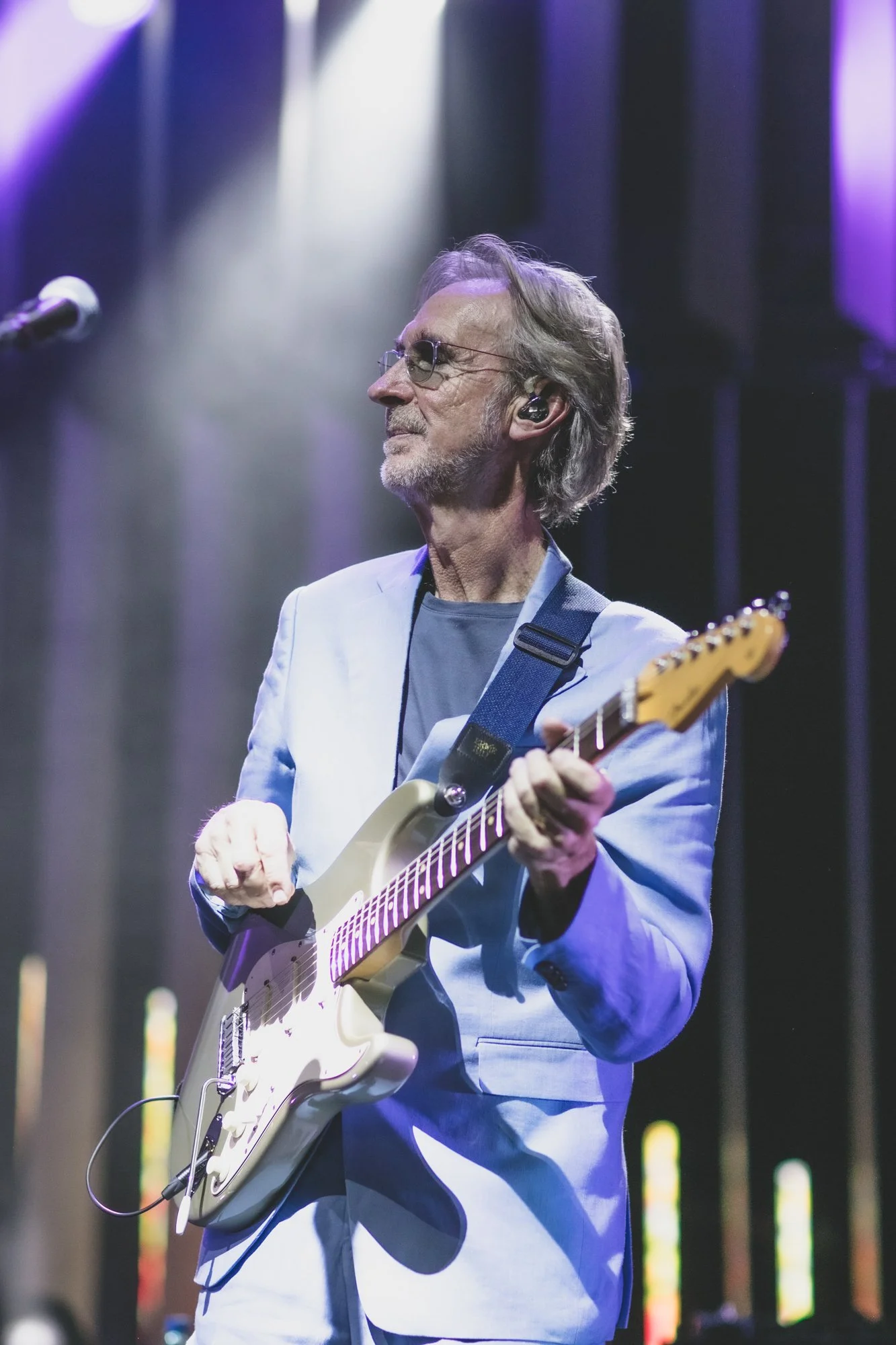A man with gray hair, wearing glasses, in a light blue suit, playing an electric guitar on stage with colorful lighting.