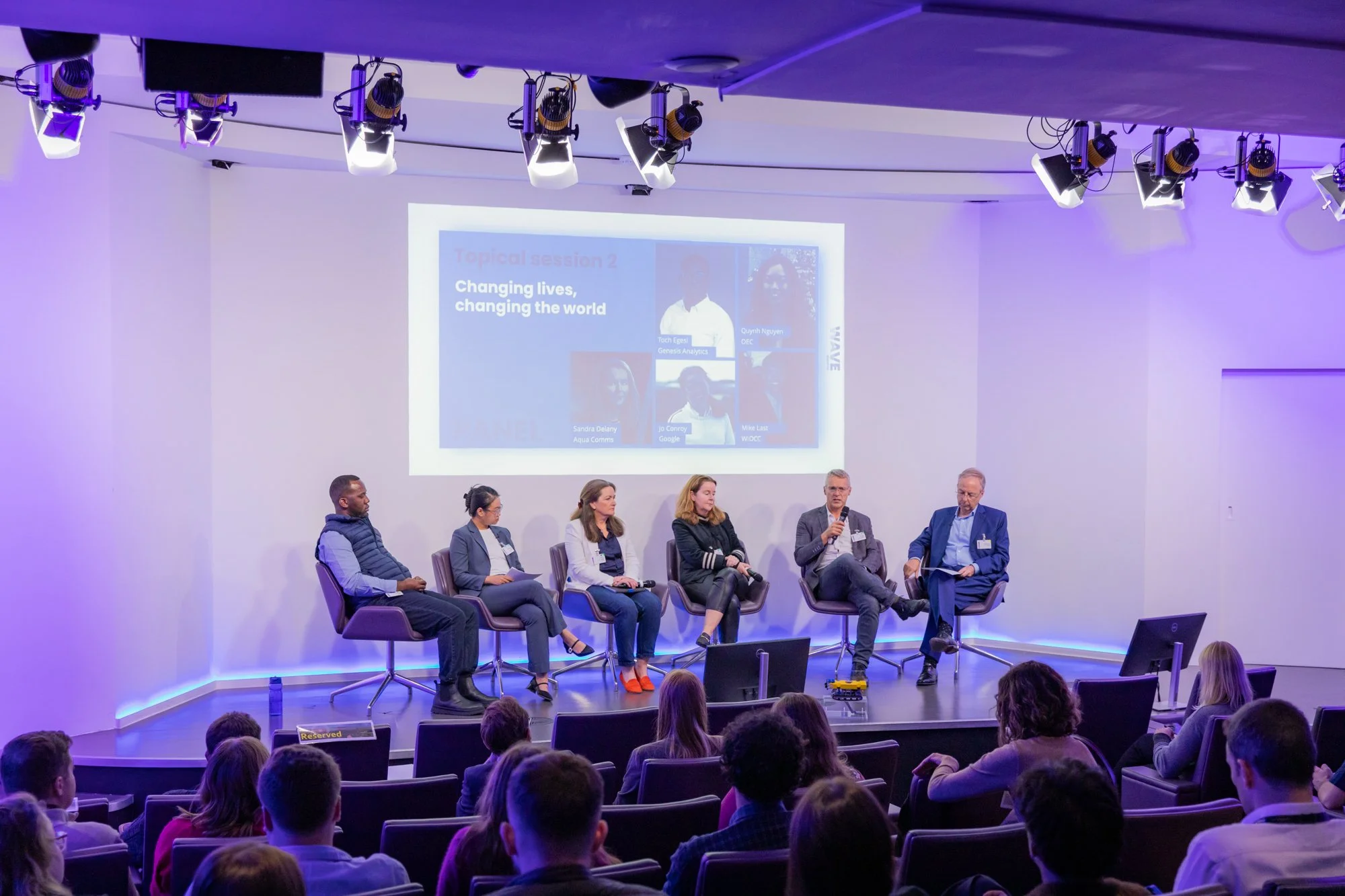 Panel discussion at a conference with six people seated on stage, audience seated in front, screen behind displaying a slide with the title 'Changing lives, changing the world' and photos of the speakers.