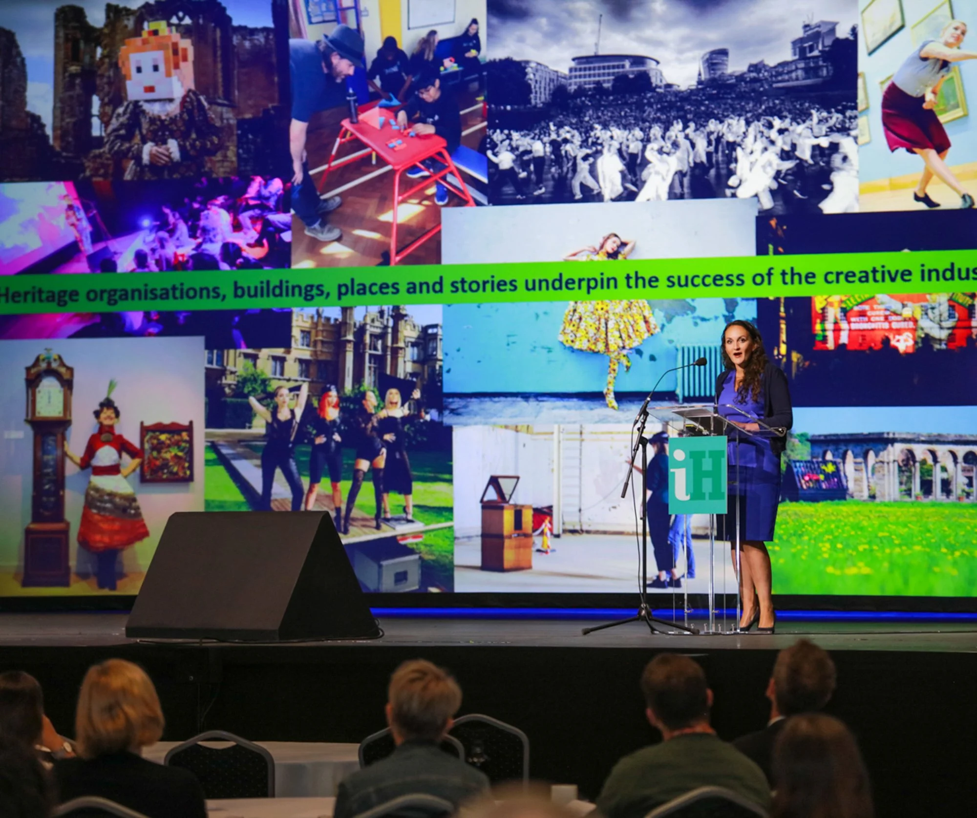 A woman giving a presentation on a stage in front of a large screen displaying various images related to culture, heritage, and creativity, with an audience seated in the foreground.