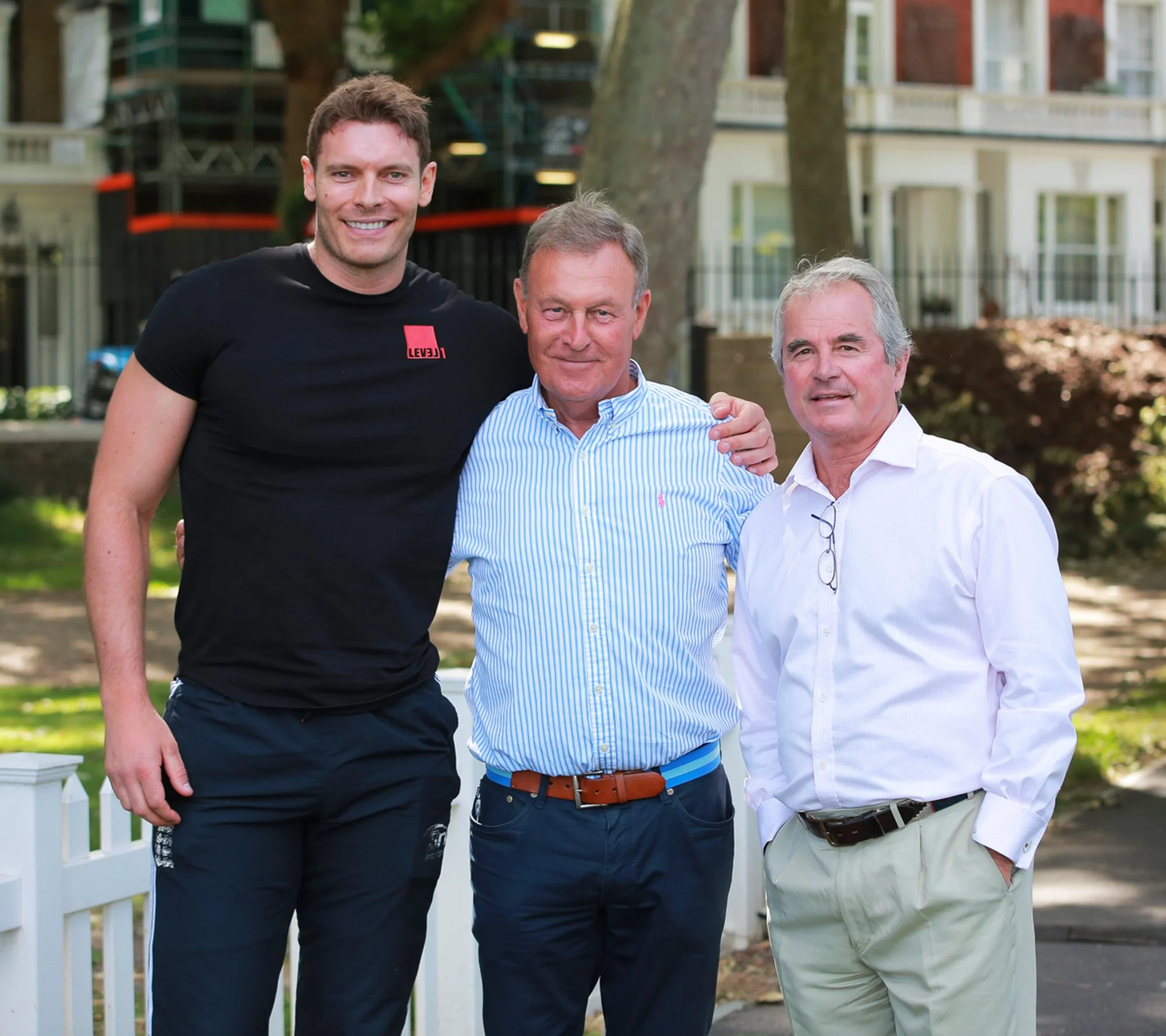 Three men standing outdoors in front of a white fence and trees, smiling for the camera.