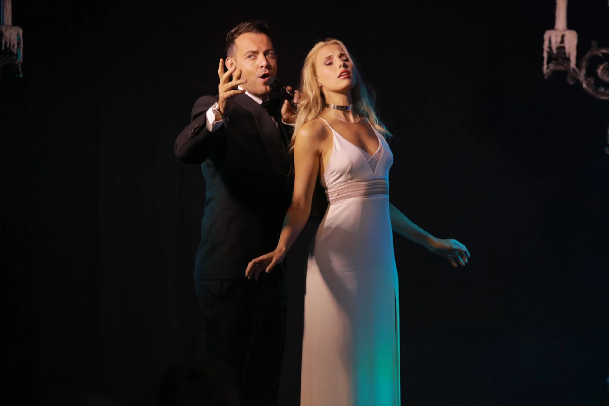 A man and woman passionately singing on stage, with the man holding a microphone and the woman in a white dress, both in dramatic poses against a dark background.