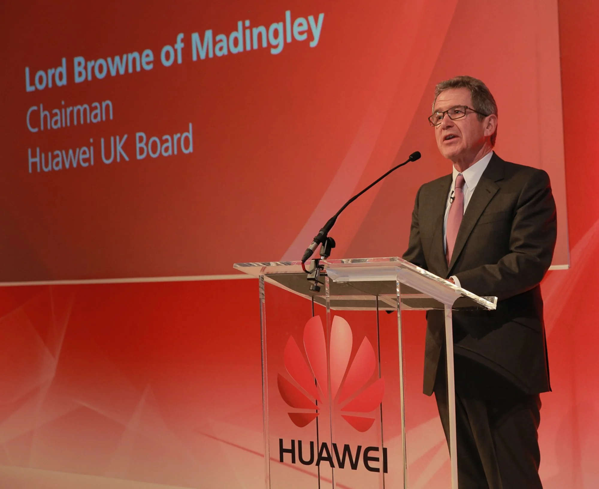 A man in a suit and glasses is speaking at a podium with a microphone. A large red screen behind him displays the text 'Lord Browne of Madingley, Chairman, Huawei UK Board.' The podium has a transparent top and a Huawei logo on the front.