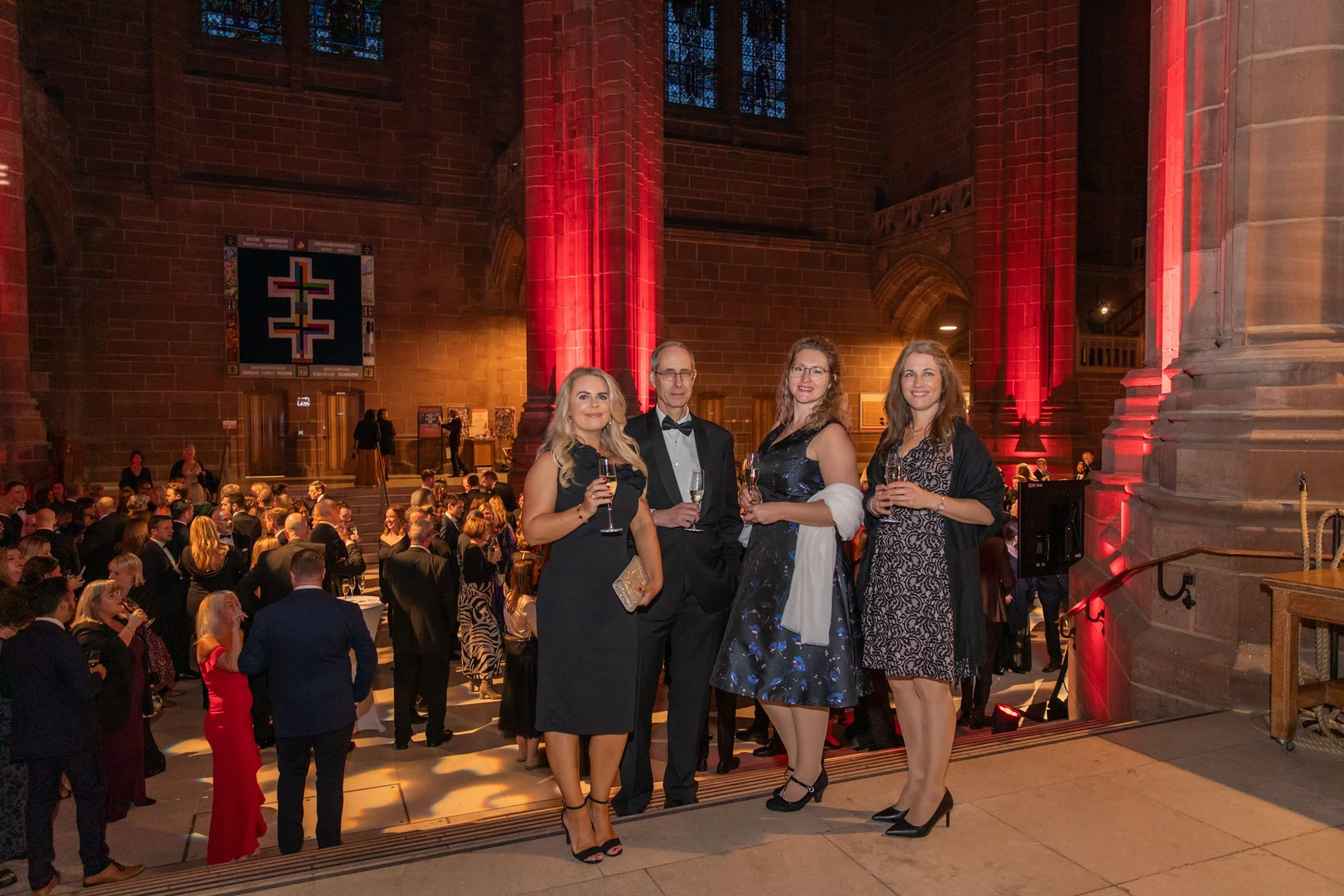 Four people dressed in formal attire standing in front of a large crowd at a church event, holding glasses of champagne, with stained glass windows and red lighting in the background.