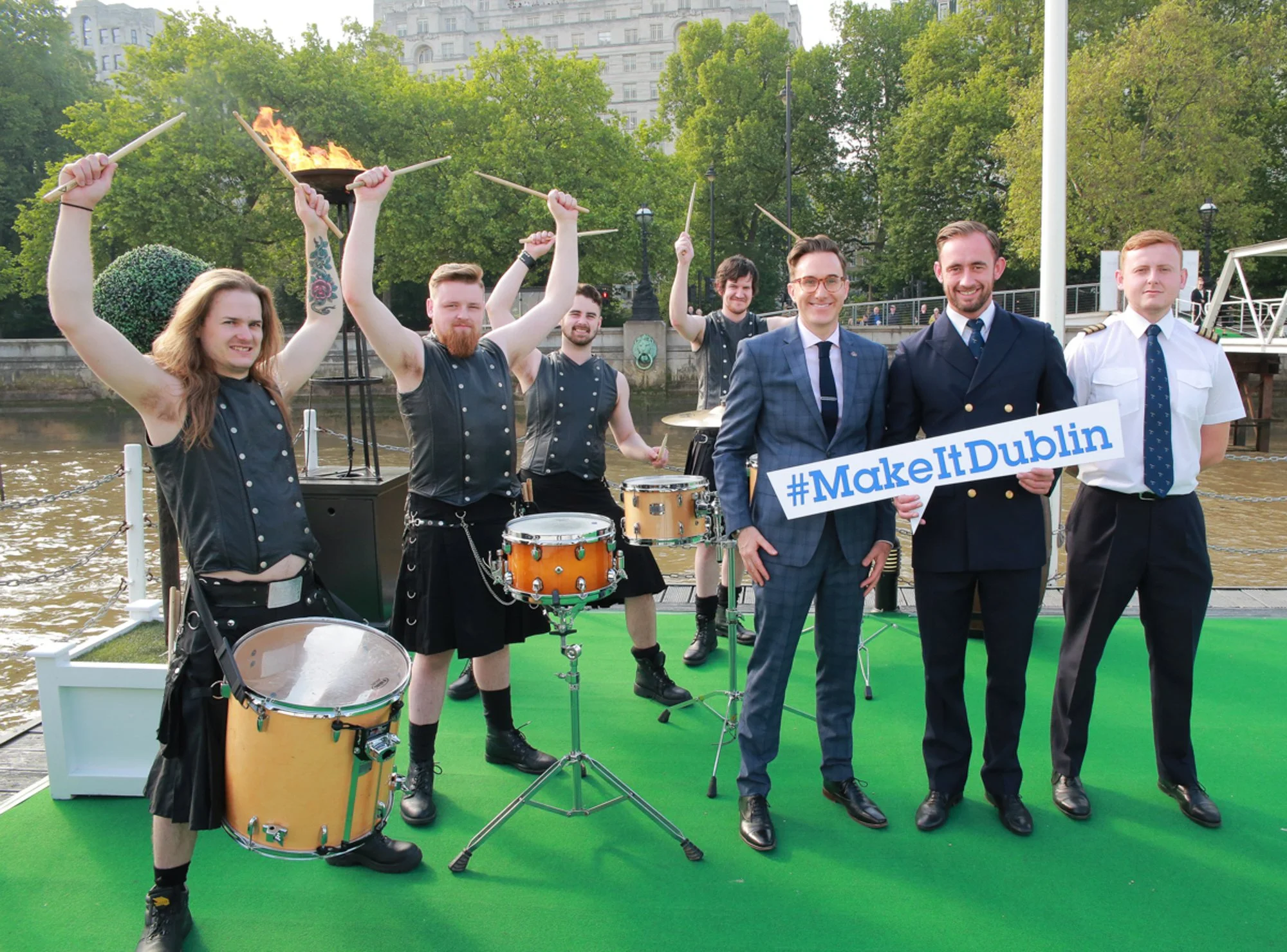 Group of seven men, some in kilts, on a green platform by a river, with trees and buildings in the background. Three men in the front hold a sign that says #MakeItDublin. Four men in the back are playing drums, with two raising drumsticks.