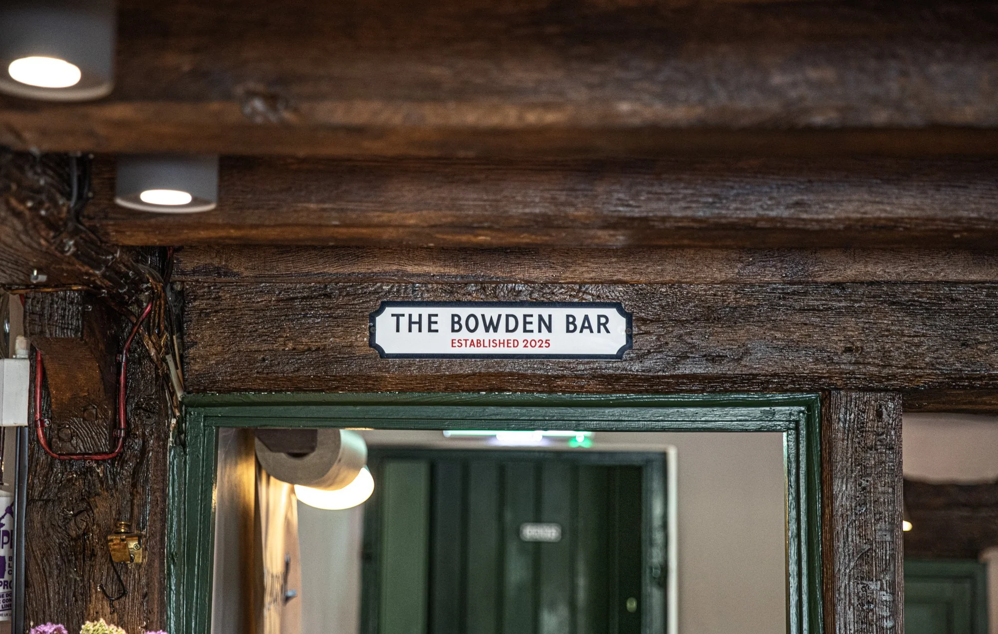 Sign reading 'The Bowden Bar' with 'Established 2025' underneath, mounted above a green door frame in a wooden interior.
