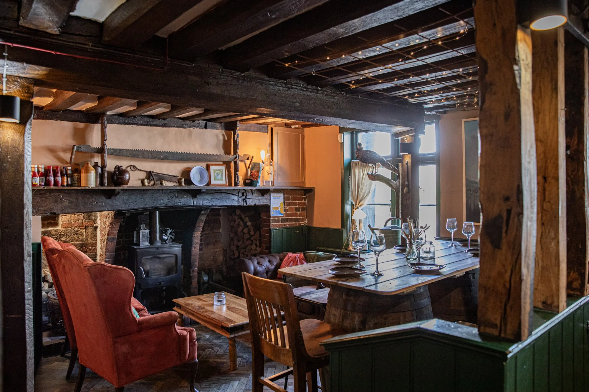 Cozy rustic interior of a cafe or restaurant with wooden beams, a wood-burning stove, a fireplace, and a long table set with wine glasses and plates, decorated with vintage and vintage-inspired decor.