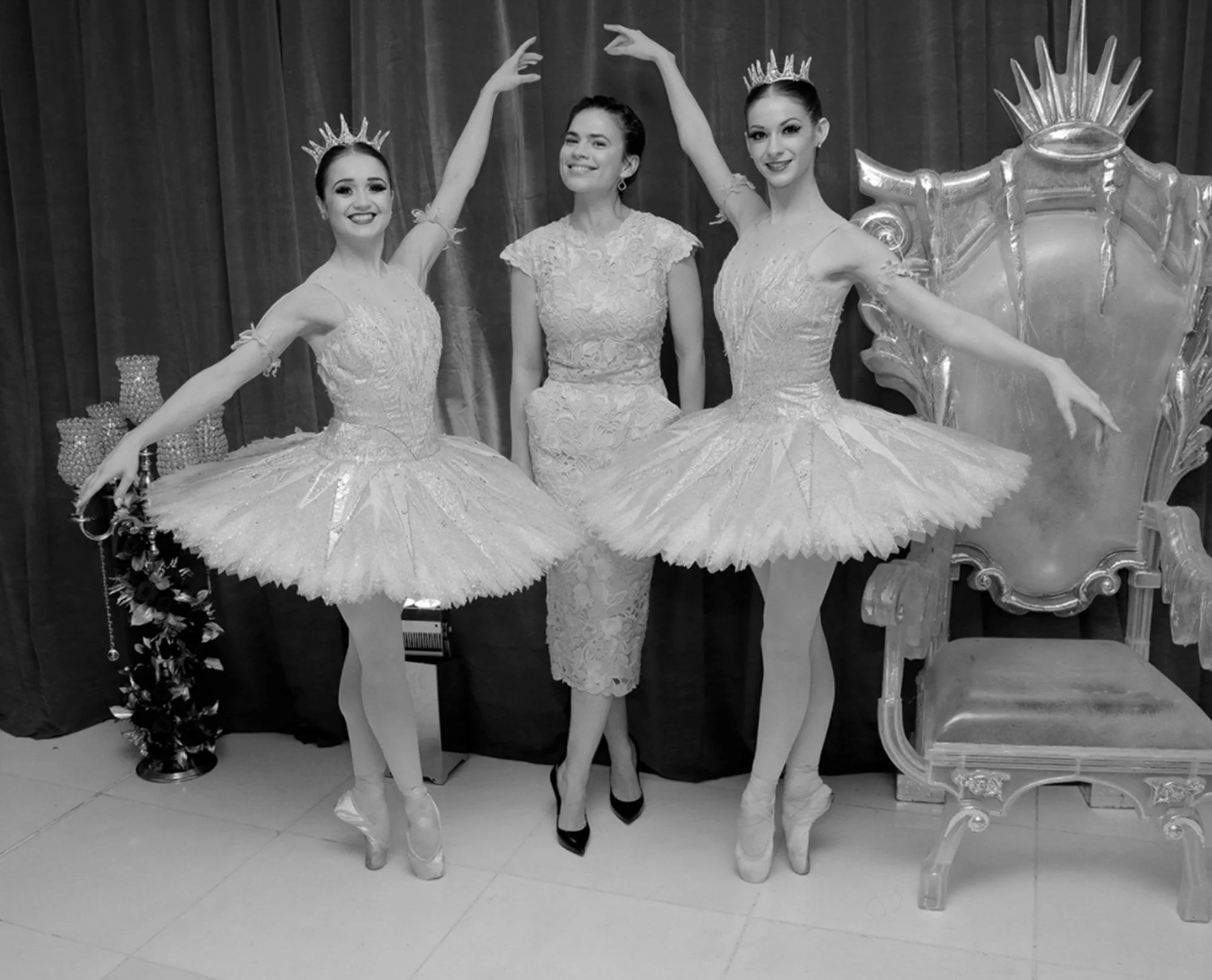 Three women in ballet costumes stand with a woman in a lace dress, all smiling. The women in ballet costumes wear tiaras and pointe shoes, with two in ballet poses, set against a dark curtain backdrop.