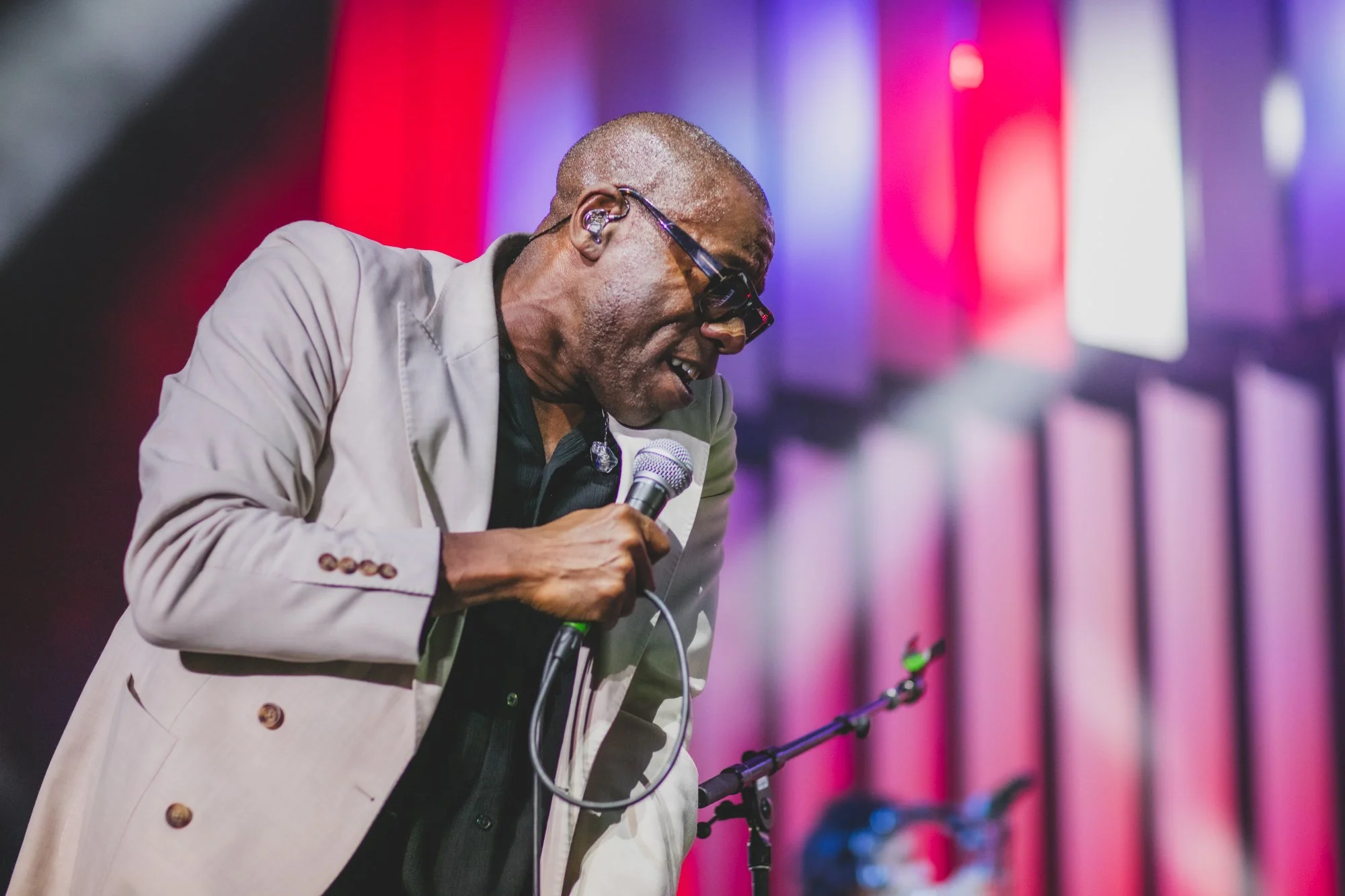 An elderly man wearing glasses and a beige suit jacket, holding a microphone, leaning forward with eyes closed, on a stage with colorful background lights.