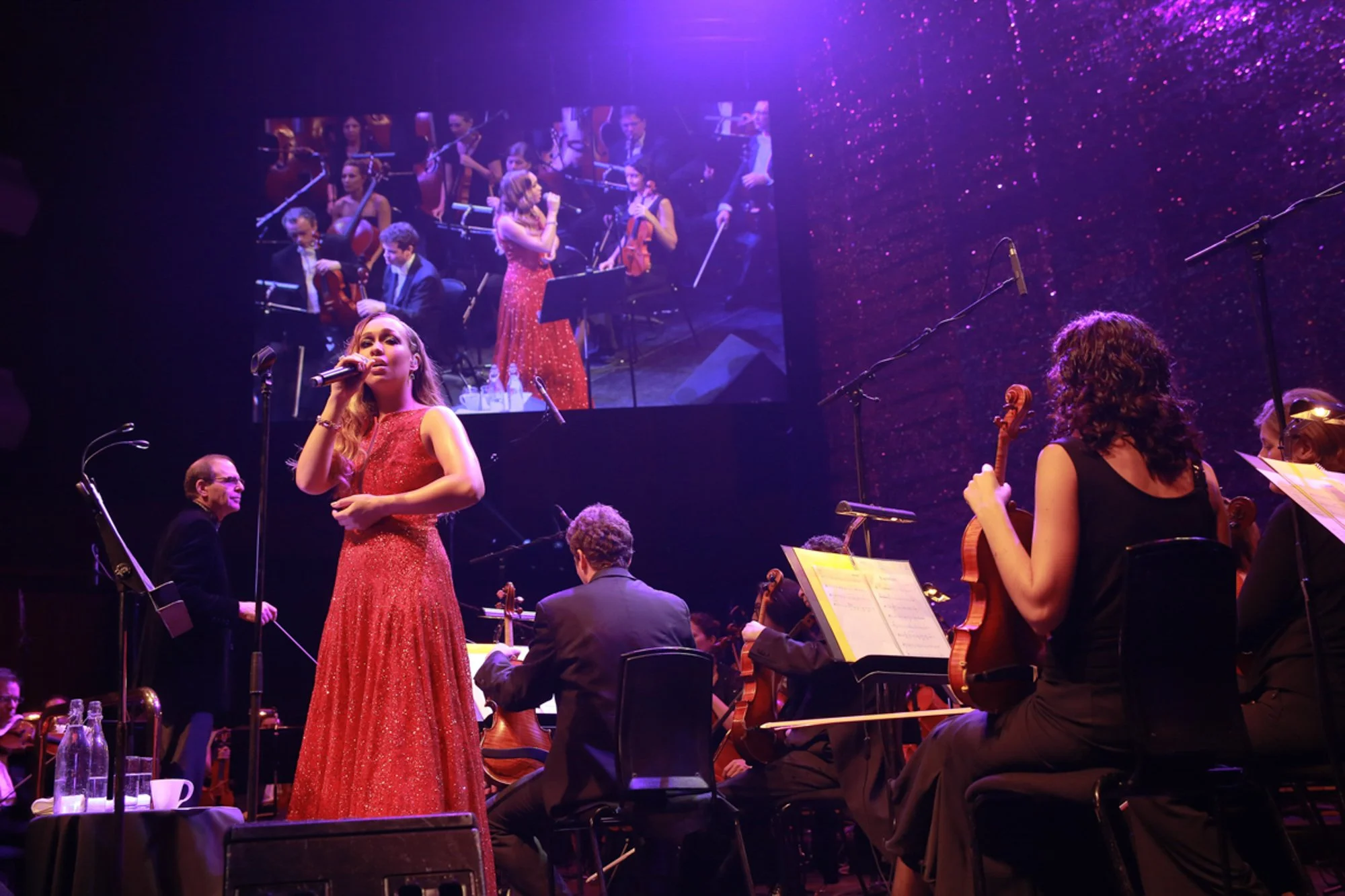 A woman in a sparkly red gown sings into a microphone on stage, with an orchestra playing behind her. A large screen behind displays a close-up of her performance, with other orchestra members and a conductor visible.