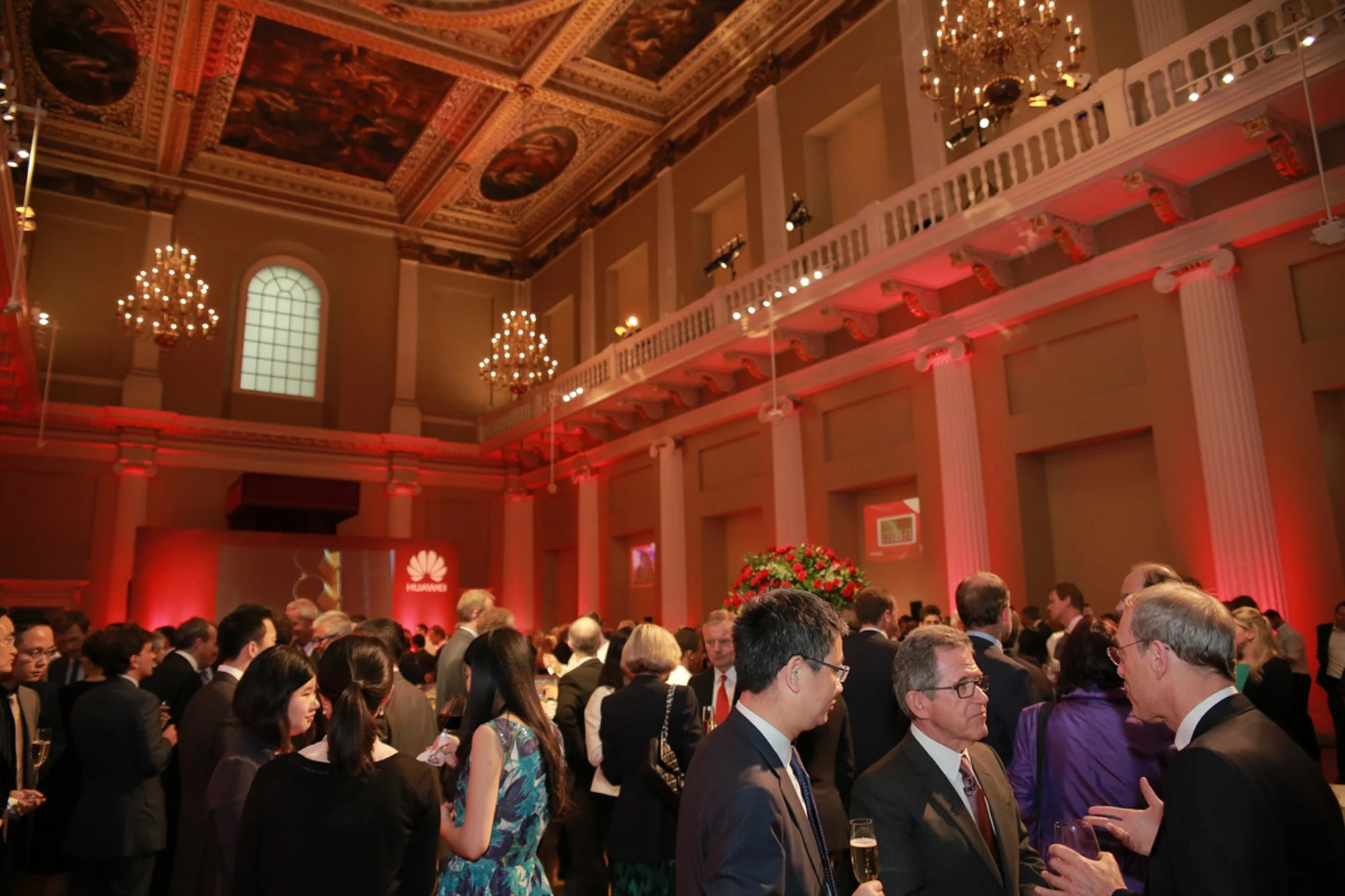 People gathered at a formal event in a grand hall with ornate ceiling, chandeliers, and red lighting, with some holding glasses of wine and engaging in conversation.