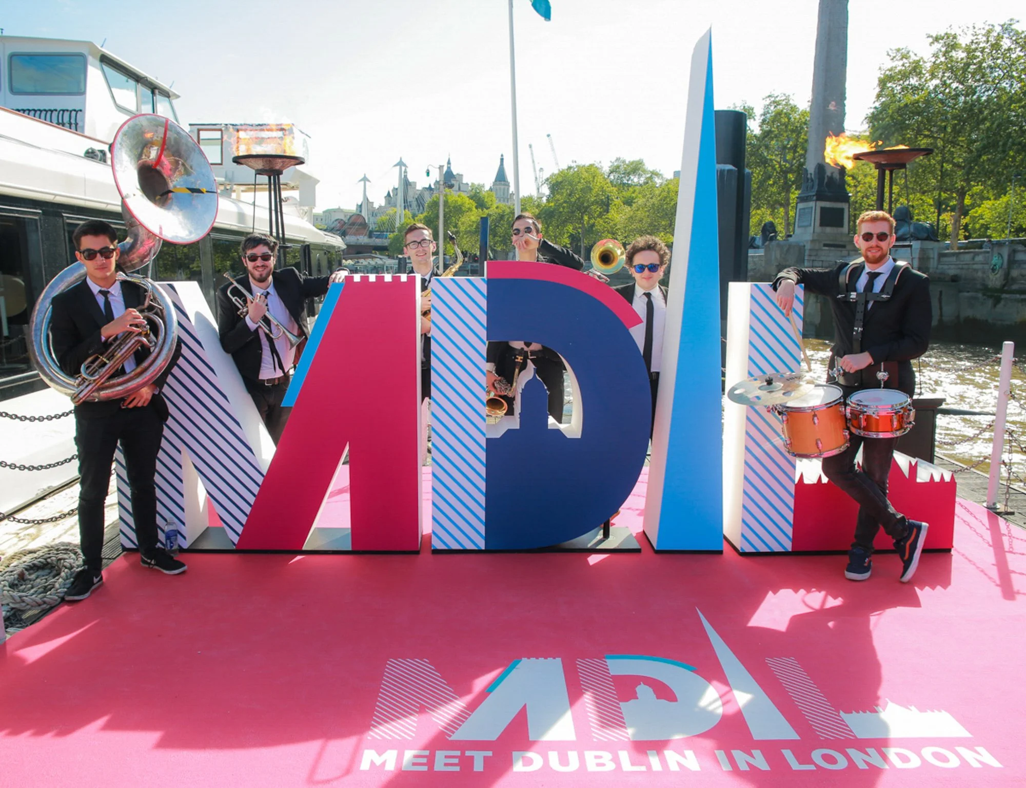 Group of six men in suits playing musical instruments on a pink stage near a river, with large colorful letters spelling 'VIRTUAL' and text below saying 'Meet Dublin in London', with trees, a statue, and buildings in the background.