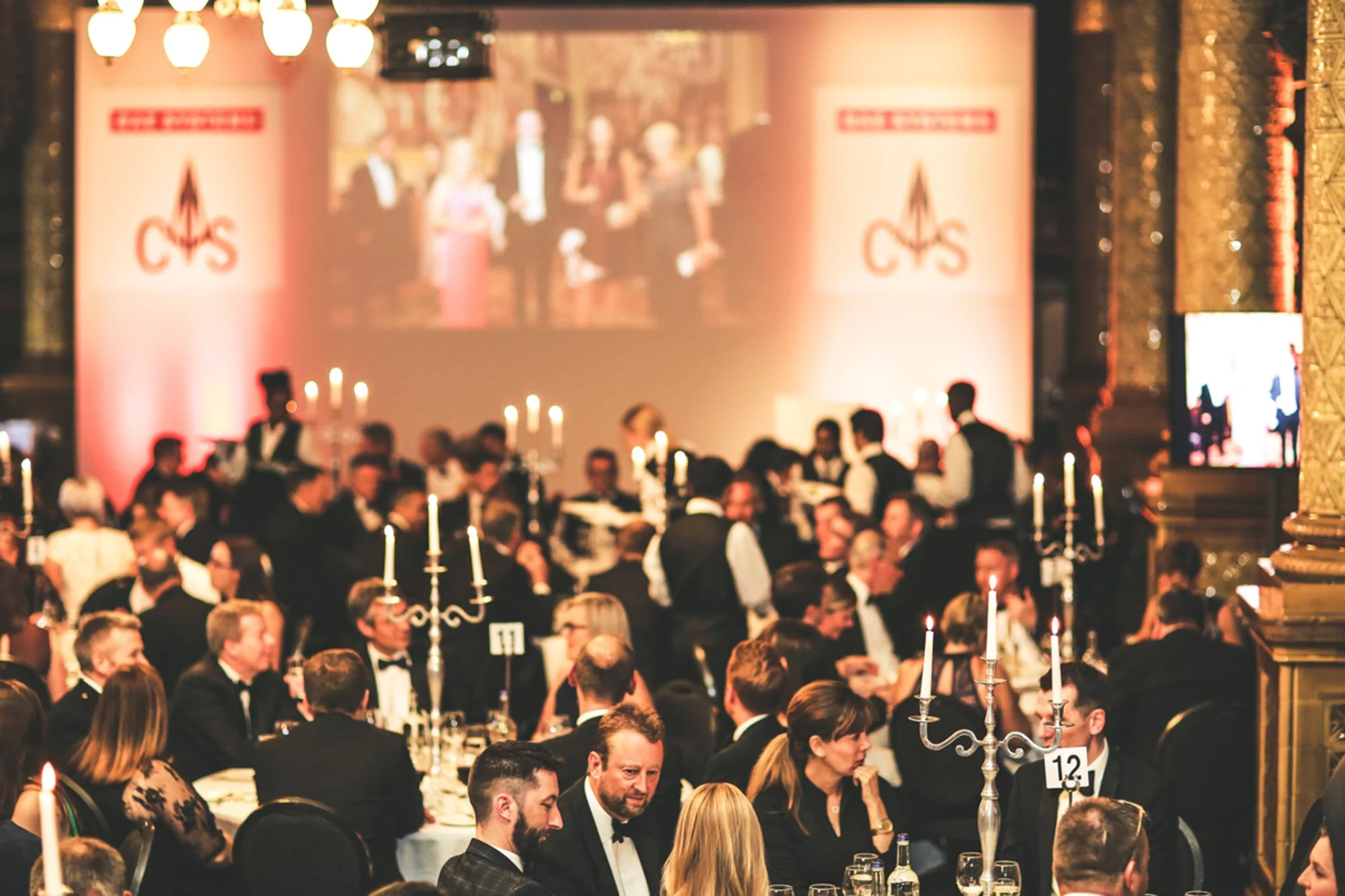 A formal banquet event with guests dressed in tuxedos and evening gowns, sitting at round tables with white tablecloths and tall candelabras, in a decorated venue with gold accents and a large screen displaying a live feed of the event.