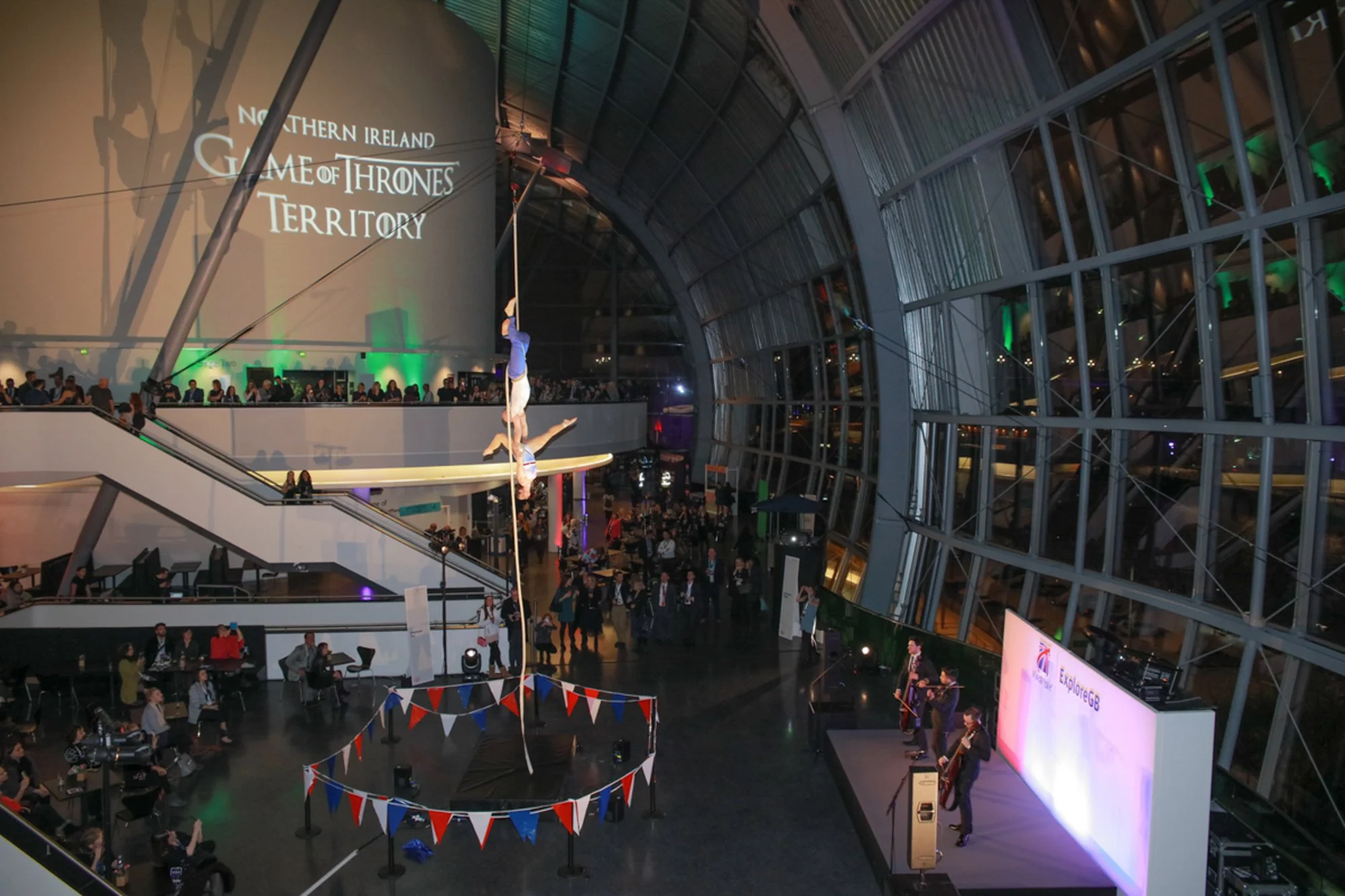 Two acrobats performing a trapeze act in a modern, glass-domed indoor venue with a crowd watching. Large screen displays 'Game of Thrones: Territory' in the background, and there is a small stage with musicians to the side.