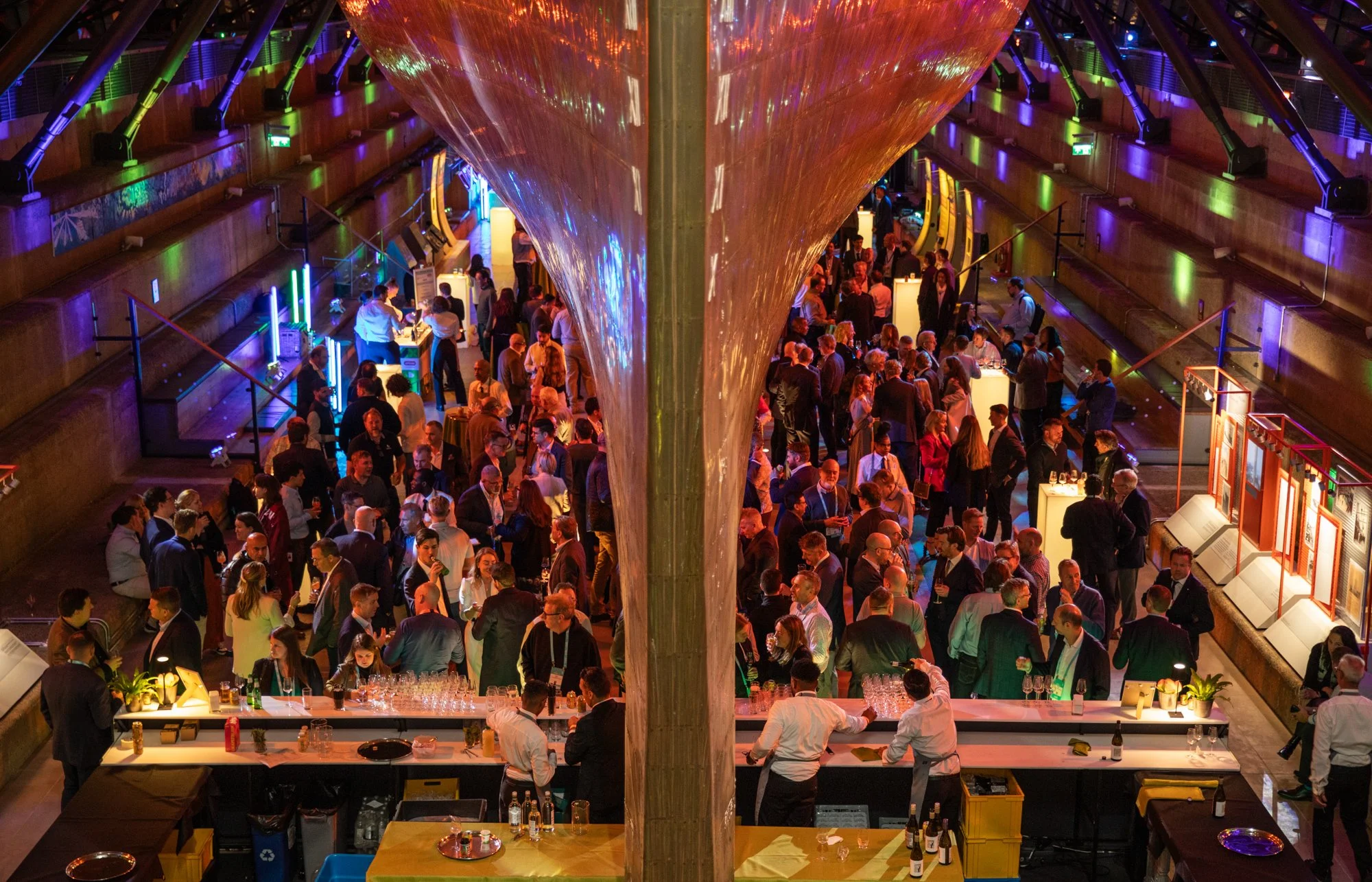 An indoor event with a large crowd of people, decorated with colorful lighting and a metallic structure in the center, possibly a concert or conference.