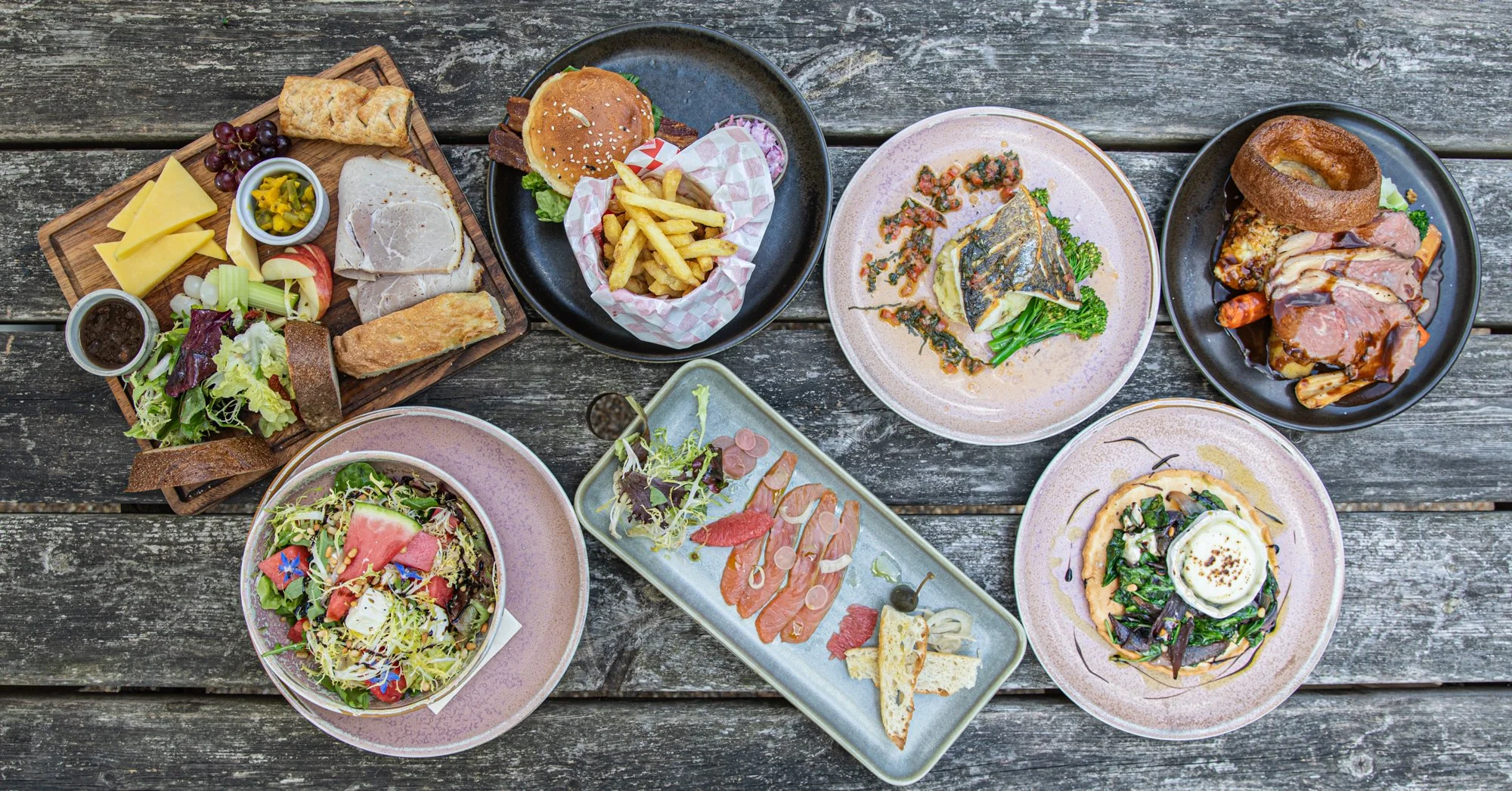 A top-down view of various plates of food on a rustic wooden table, including salads, sandwiches, a burger with fries, a plate of fish with greens, a meat dish, and an assorted charcuterie platter.