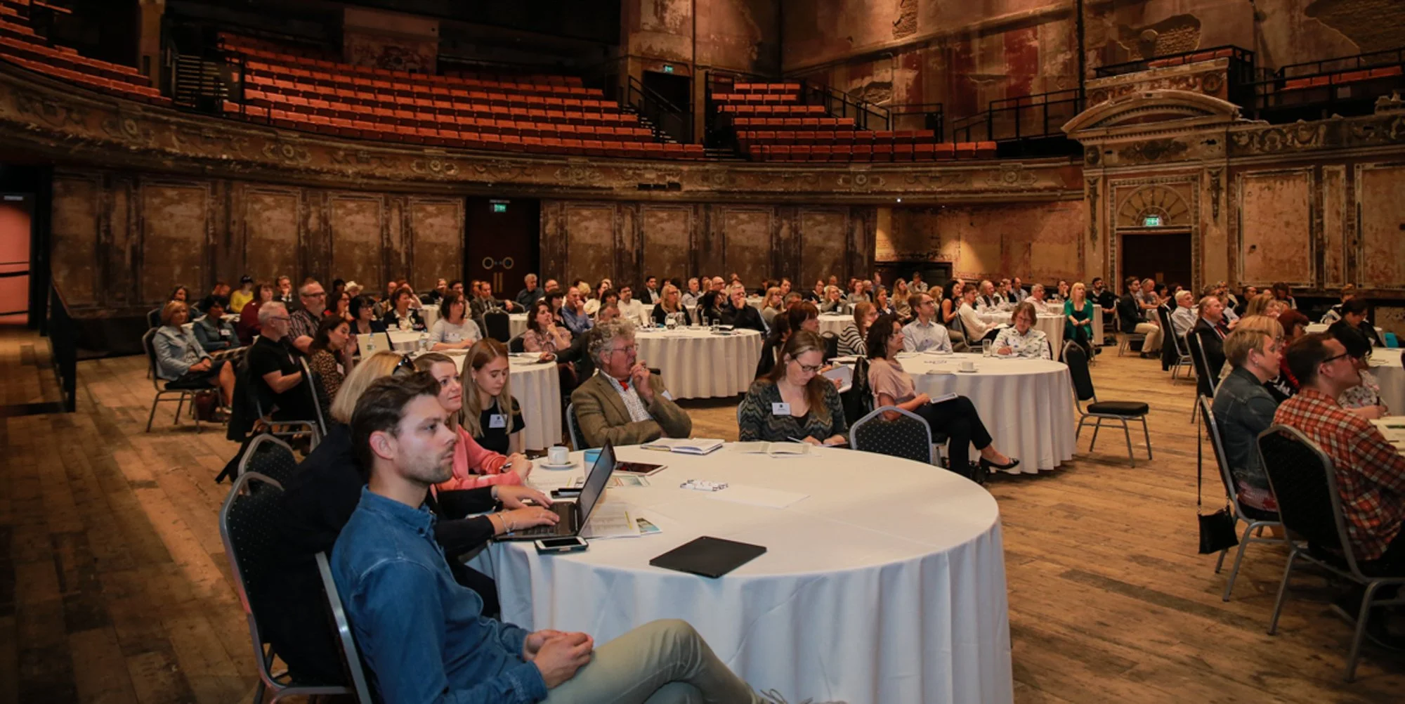 Audience attending a conference in an ornate theater with round tables and laptops, facing the stage area.
