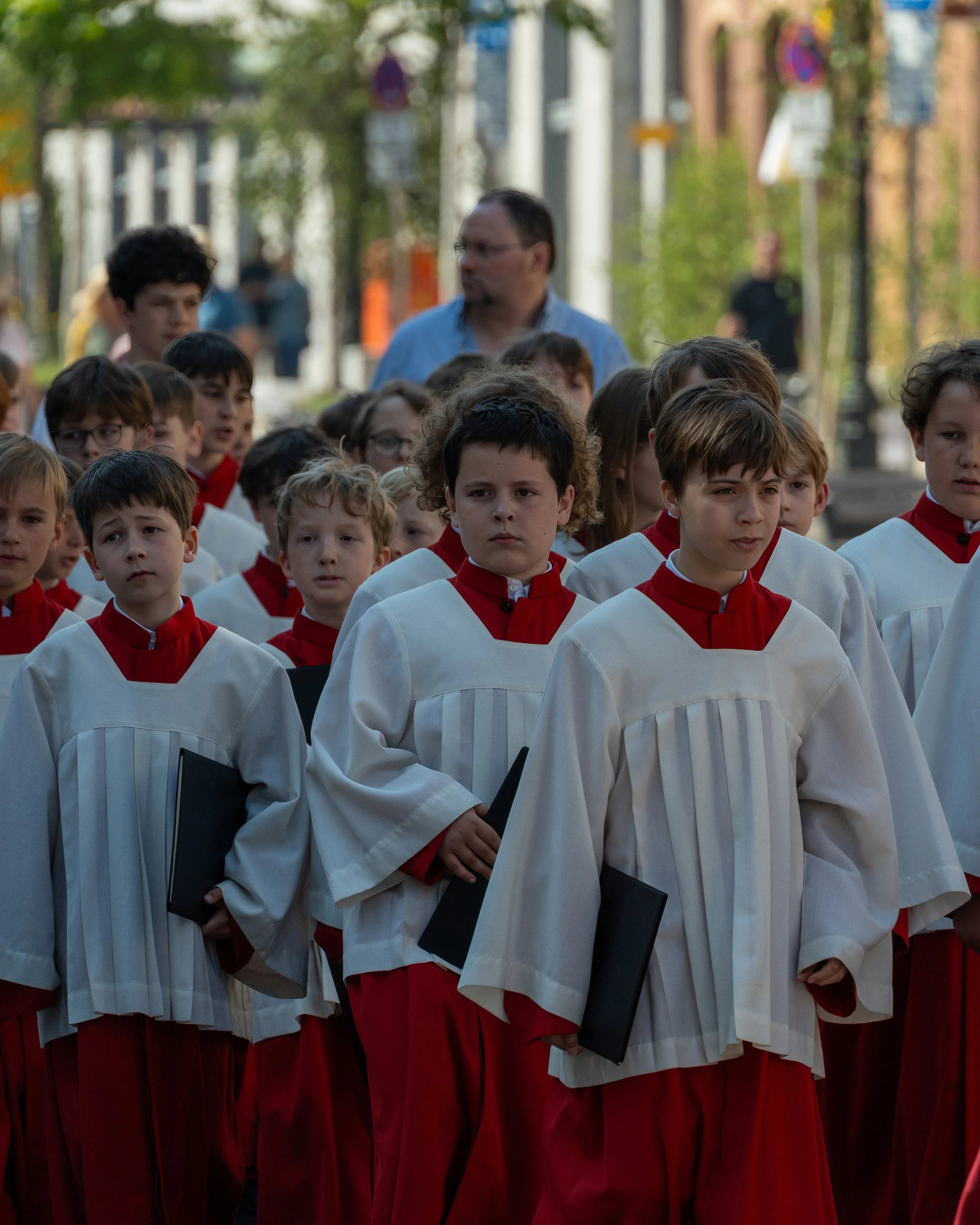 Group of children dressed in choir robes holding black folders, gathered outdoors for a ceremony, with some adults in the background.