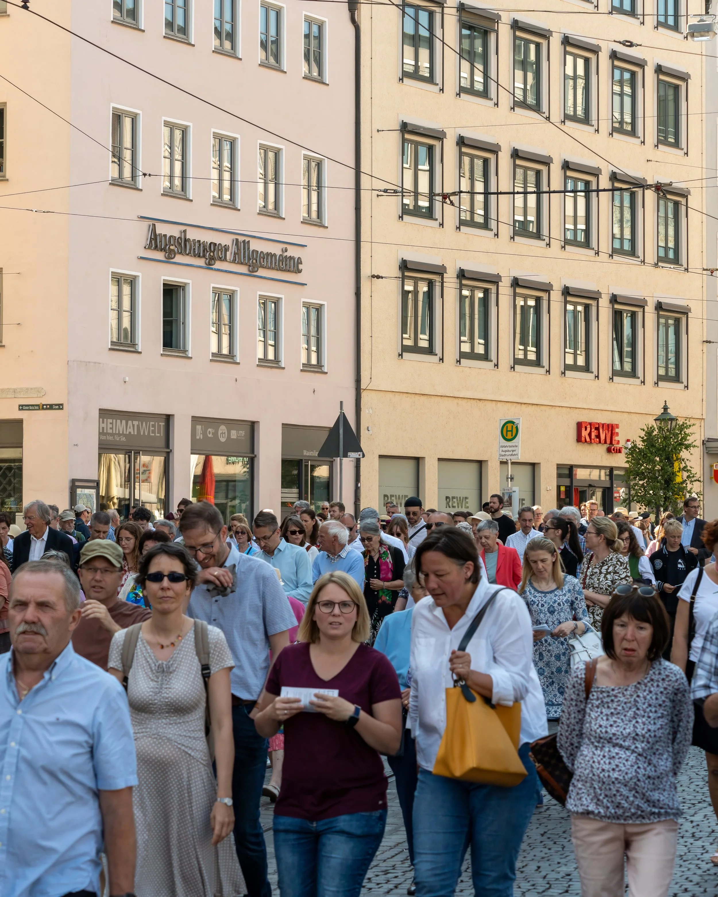 A crowd of people walking in a city street with buildings and shop signs in the background.