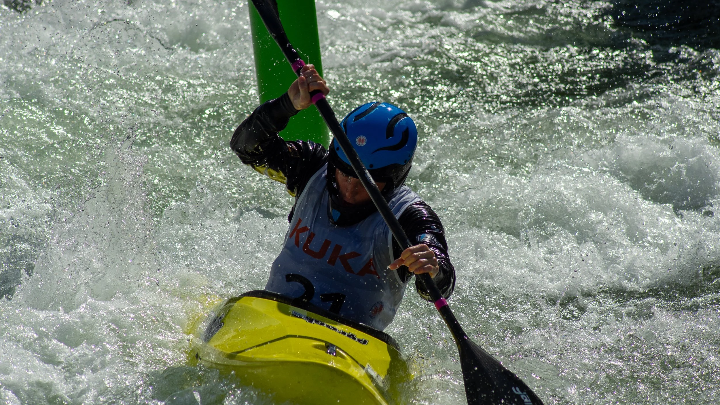 A kayaker wearing a blue helmet and black jacket navigates through rough white water in a yellow kayak.
