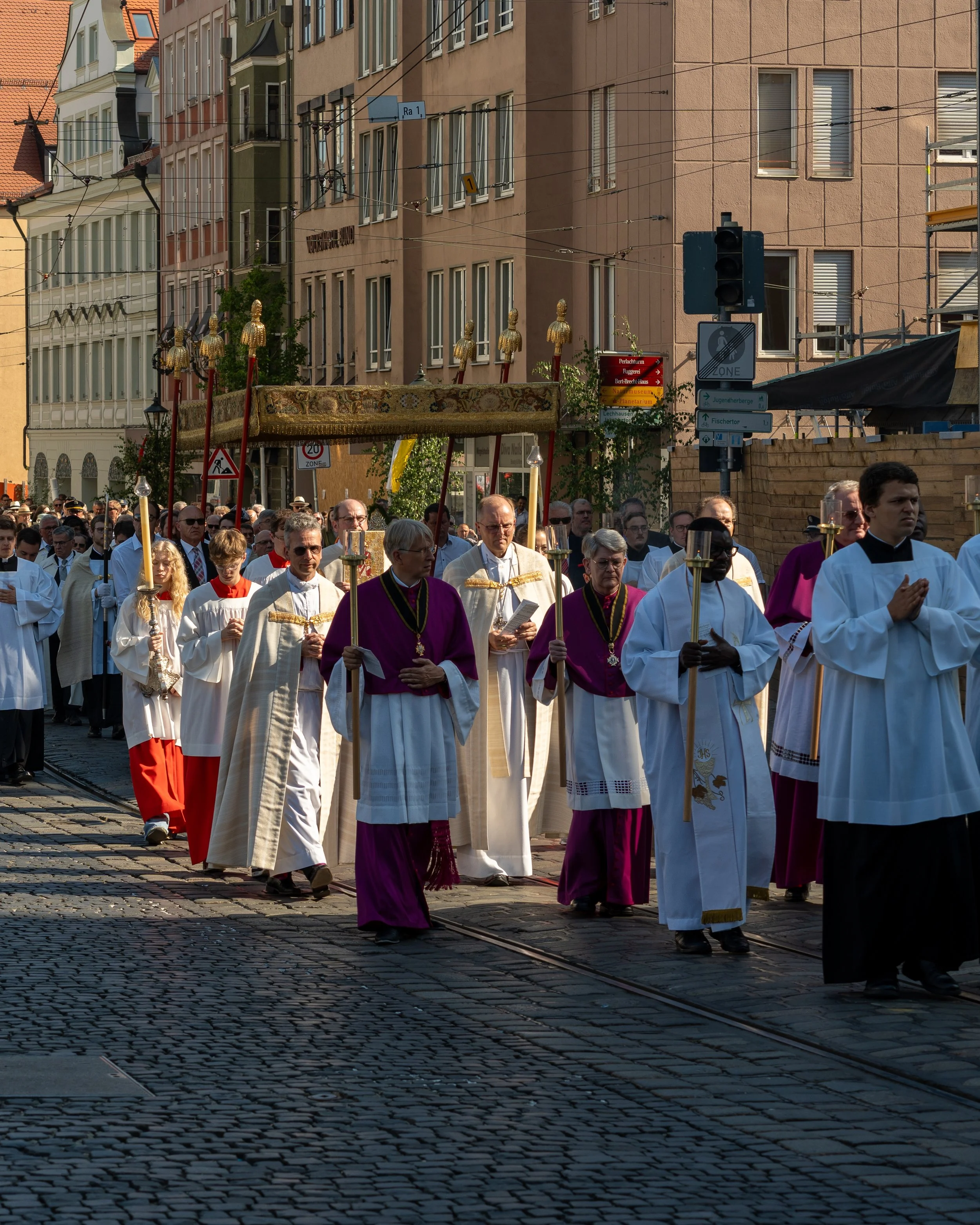 A religious procession with clergy and procession participants walking on cobblestone street, carrying a canopy, lanterns, and candles during daytime in an urban area with pink and beige buildings.