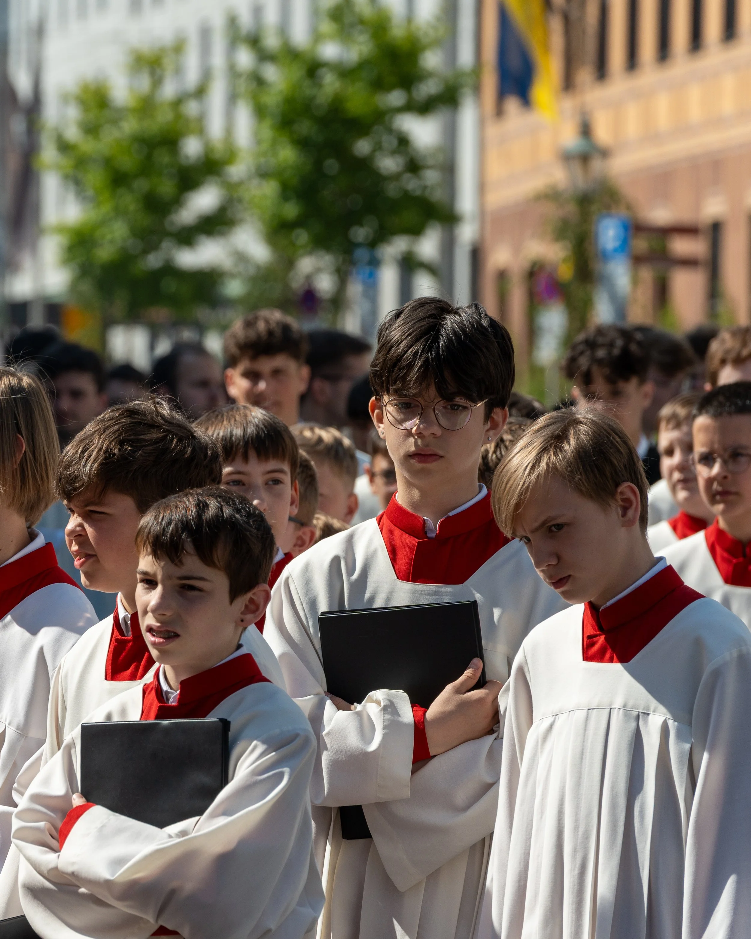 Young boys dressed in white choir robes with red collars, holding black folders, standing in formation outdoors during a church event or procession.