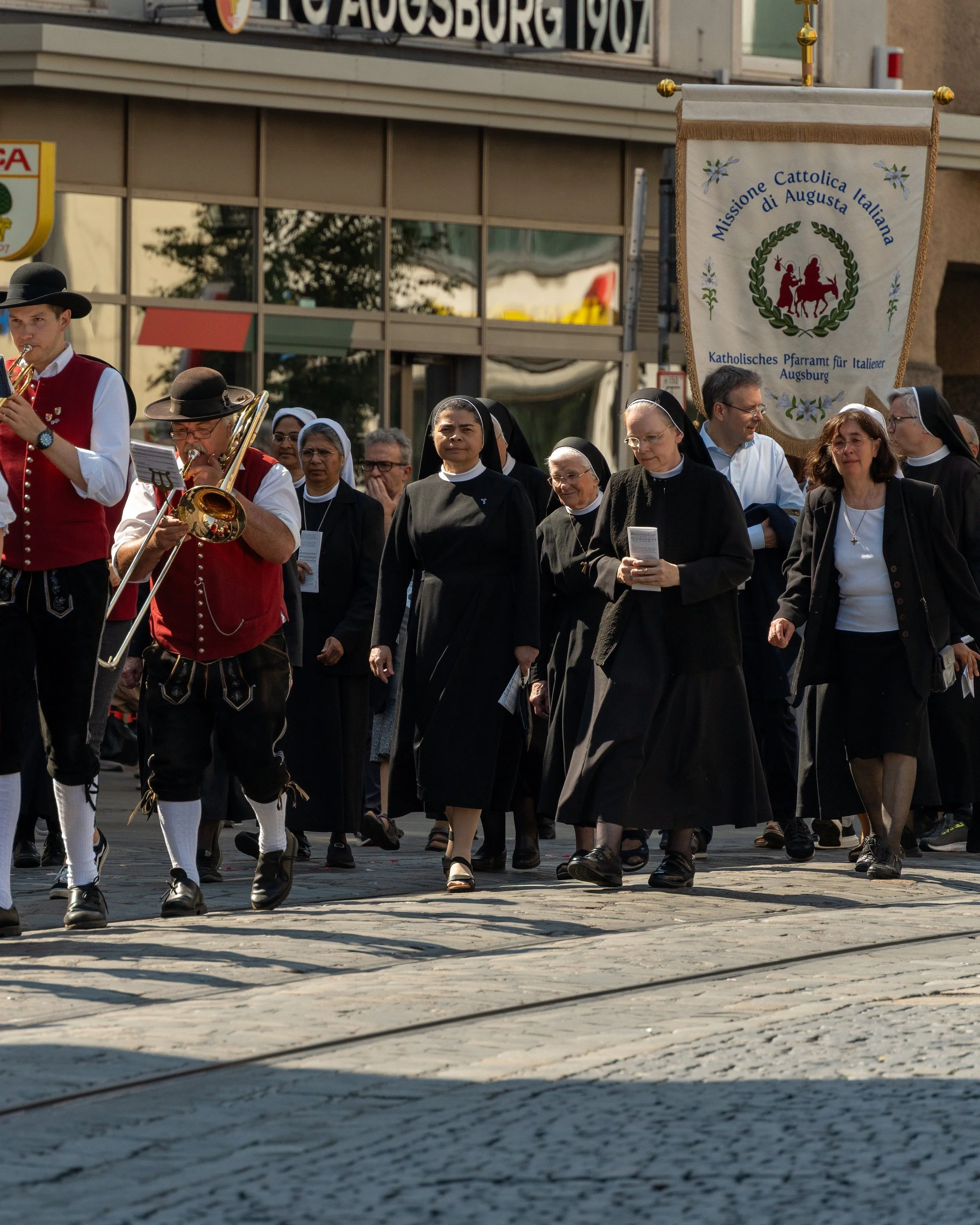 Group of people participating in a religious procession, including nuns and musicians, on a city street. A banner reads 'Missione Cattolica Italiana di Augusta' and others in the background have Italian text.