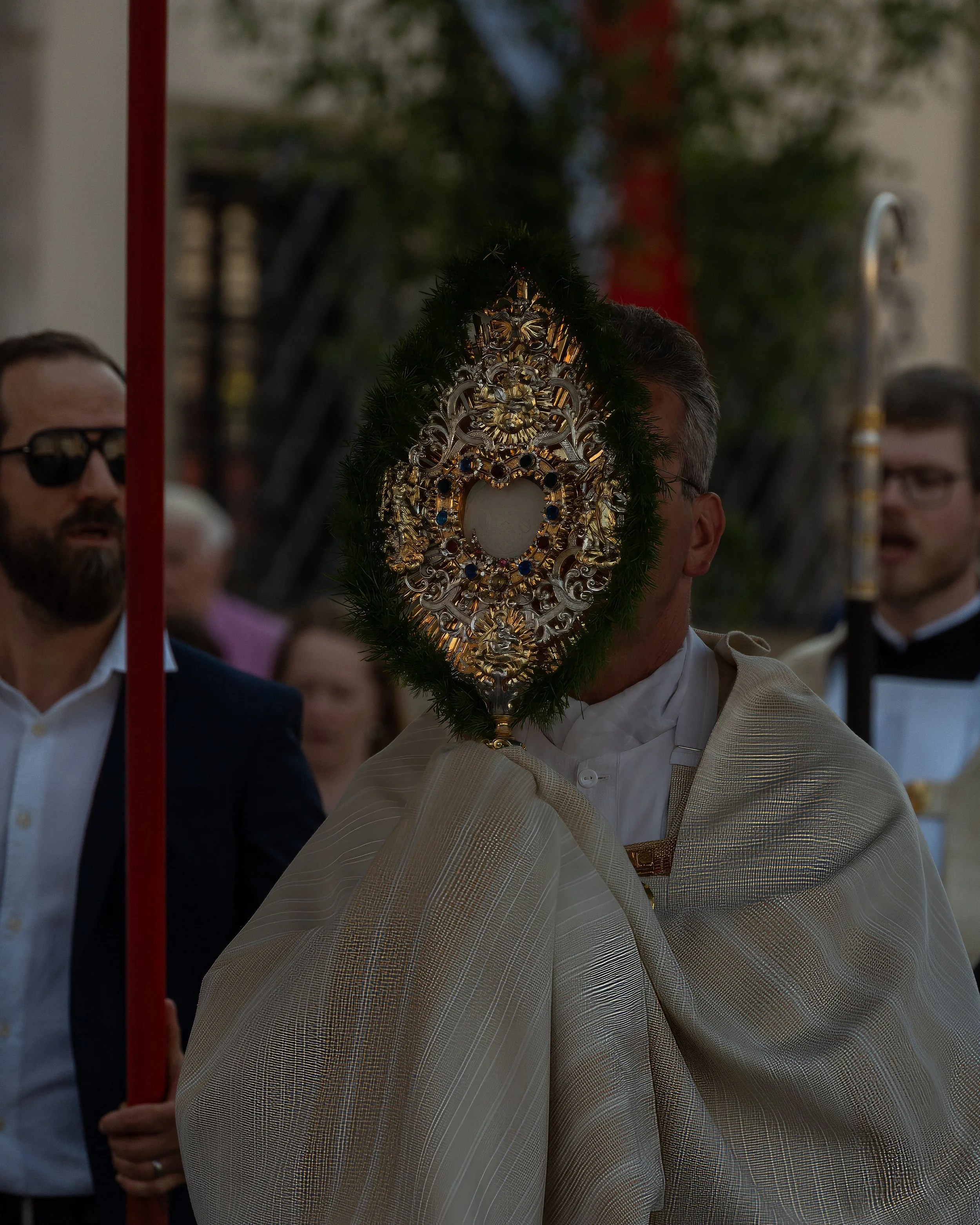 A person dressed in a white robe holding a decorative religious relic or icon with ornate gold and silver details and green foliage framing the relic face. Several other people are visible in the background at what appears to be a religious processio