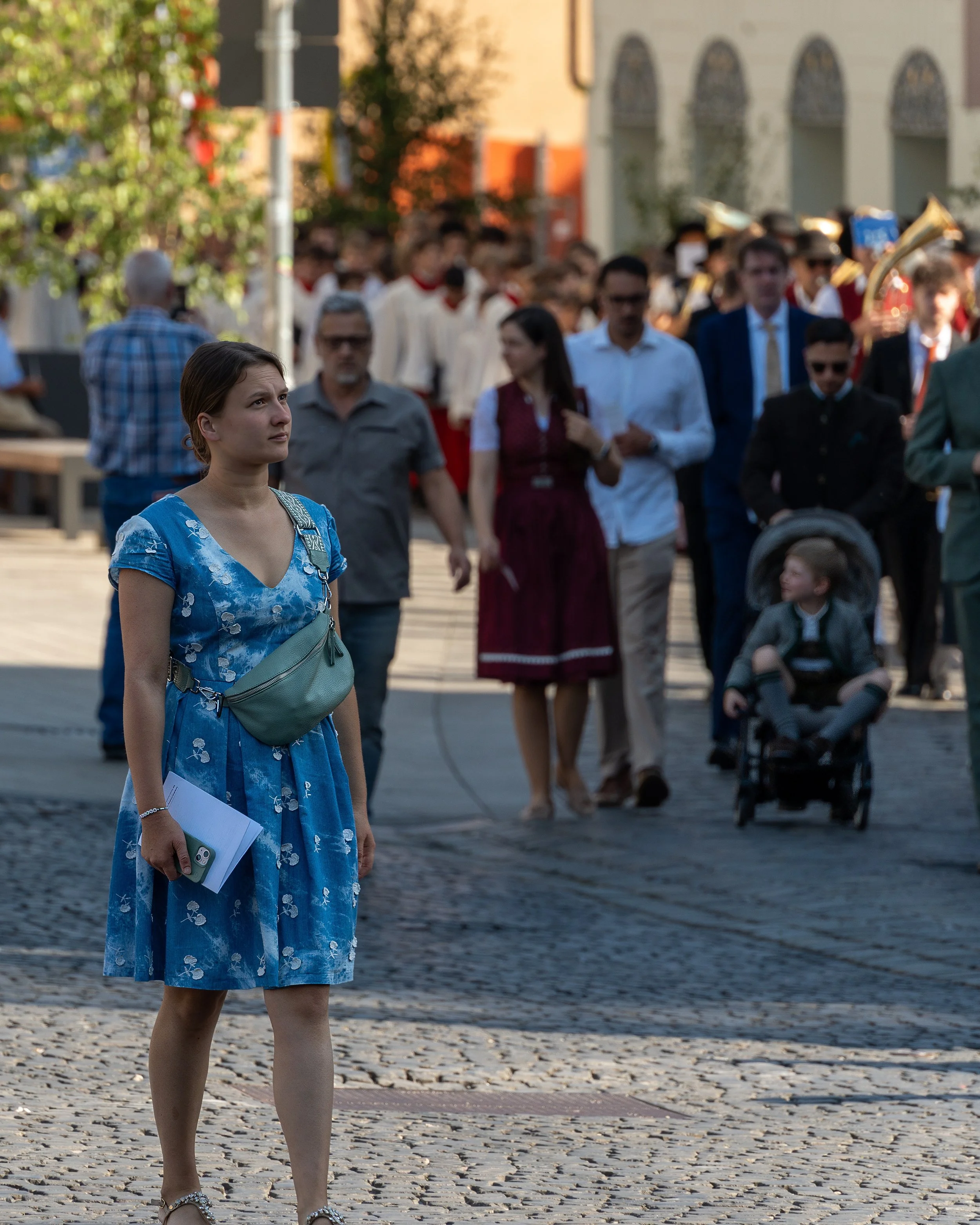 A young woman in a blue dress standing on a cobblestone street, holding a phone and papers, looking curiously at a parade or gathering in the background.