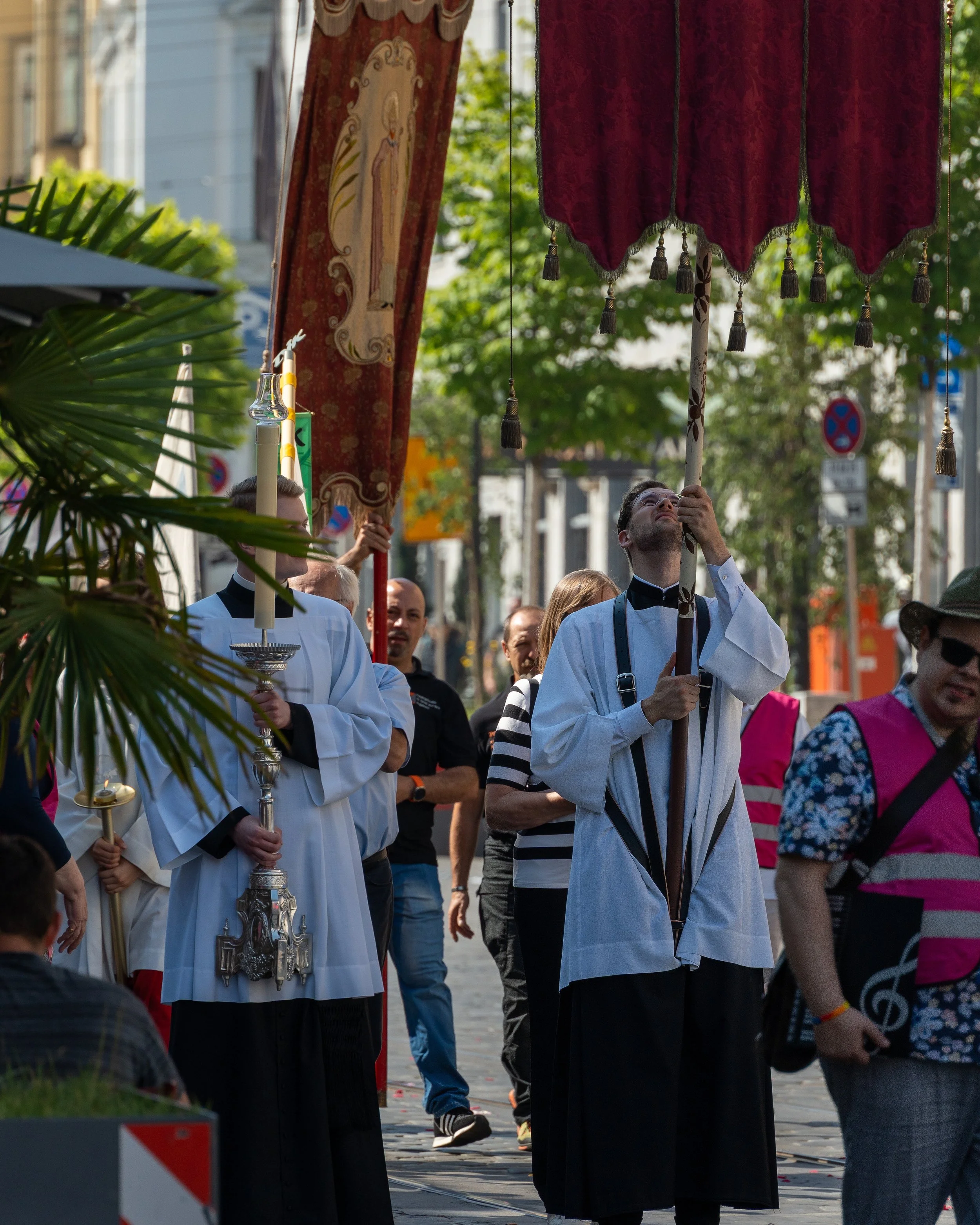 Clergy members in white robes holding a large ornate canopy during a street procession with onlookers nearby.
