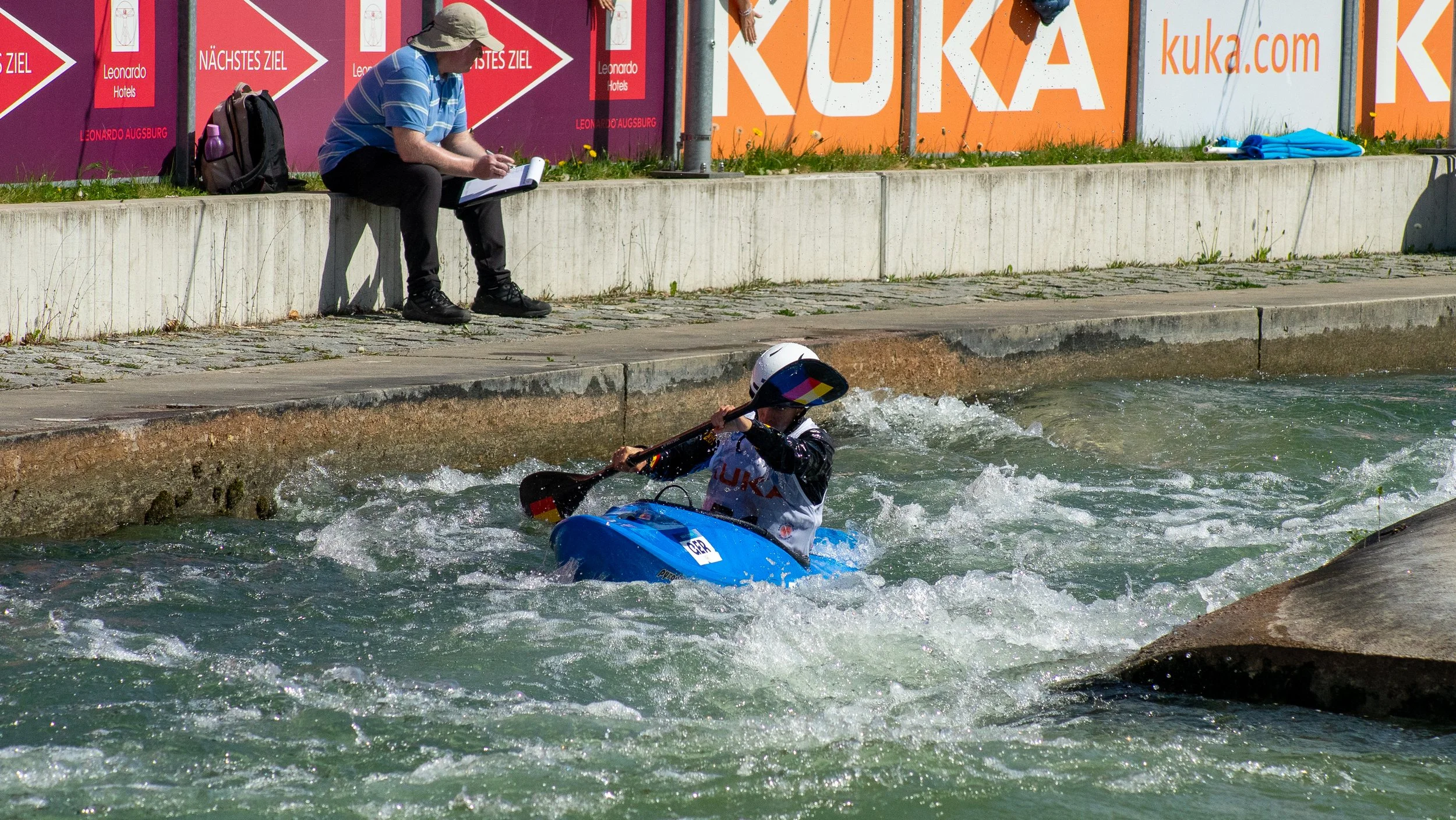 A kayaker wearing a helmet and a life vest paddling in a water canal, with a person sitting on the bank taking notes, and advertising banners along the edge of the canal.