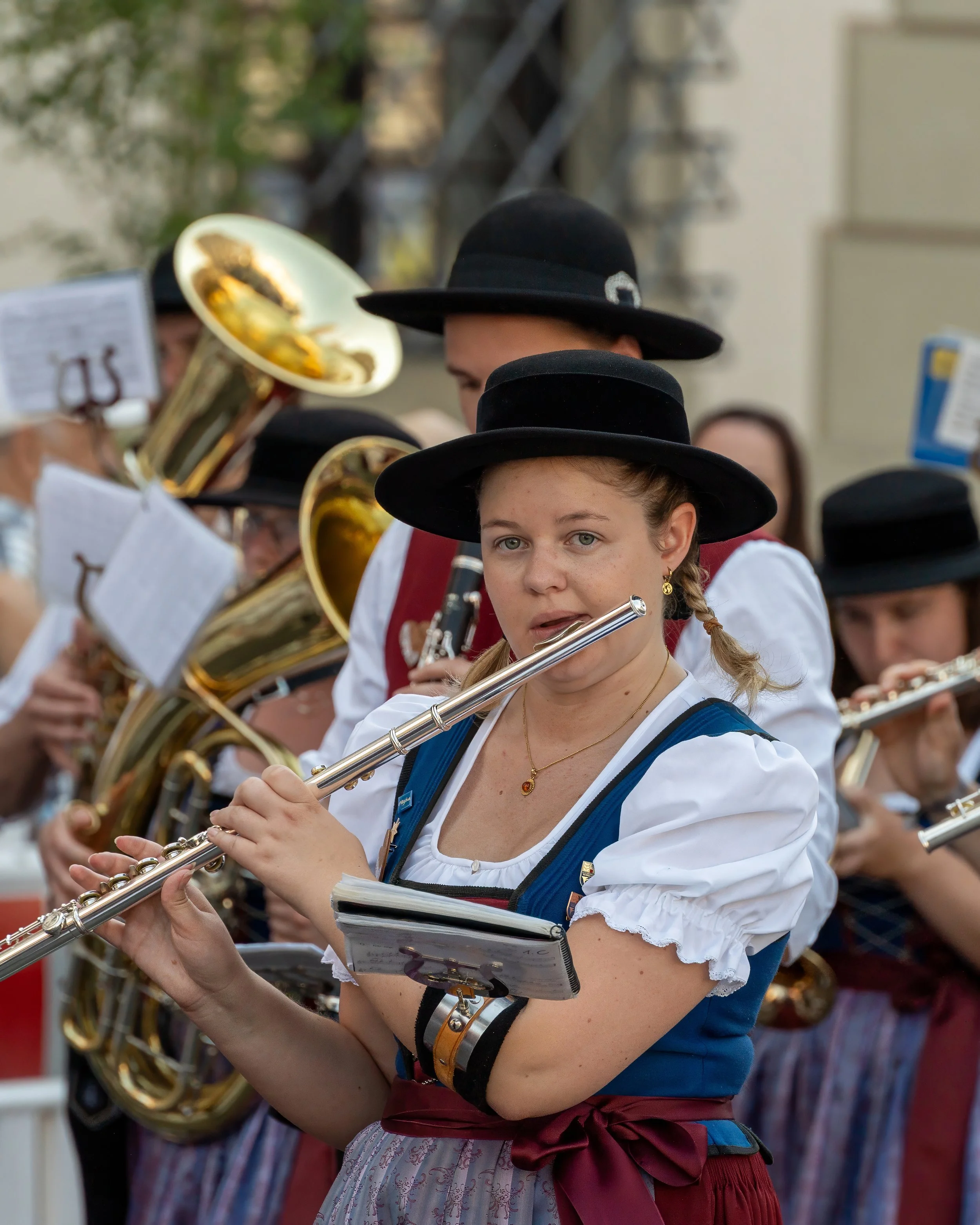 Young woman in traditional Bavarian dress playing a flute in a band with other musicians, some playing brass instruments, all wearing black hats.