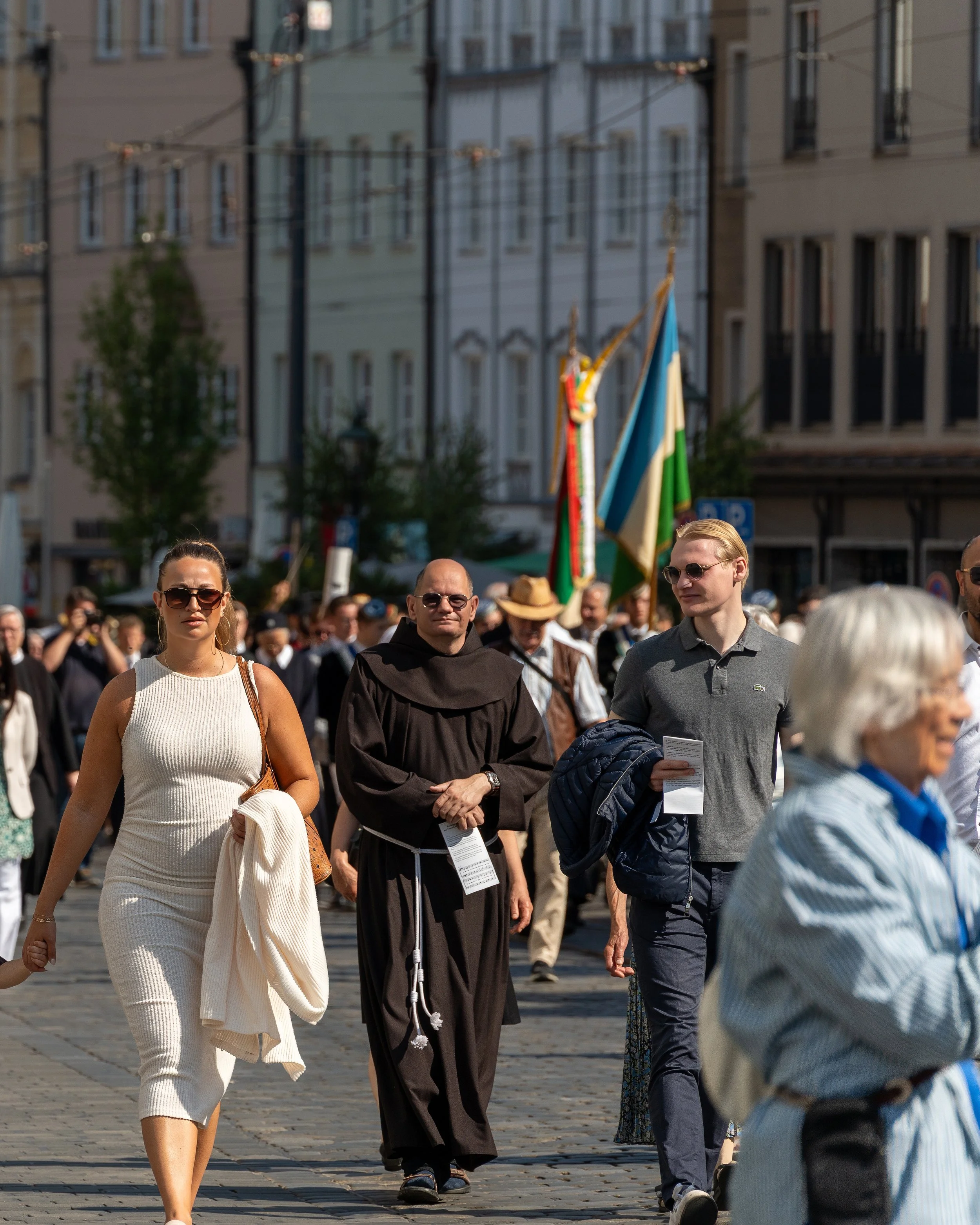 People participating in a procession or parade on a city street, with some holding flags and banners, in front of multi-story buildings.