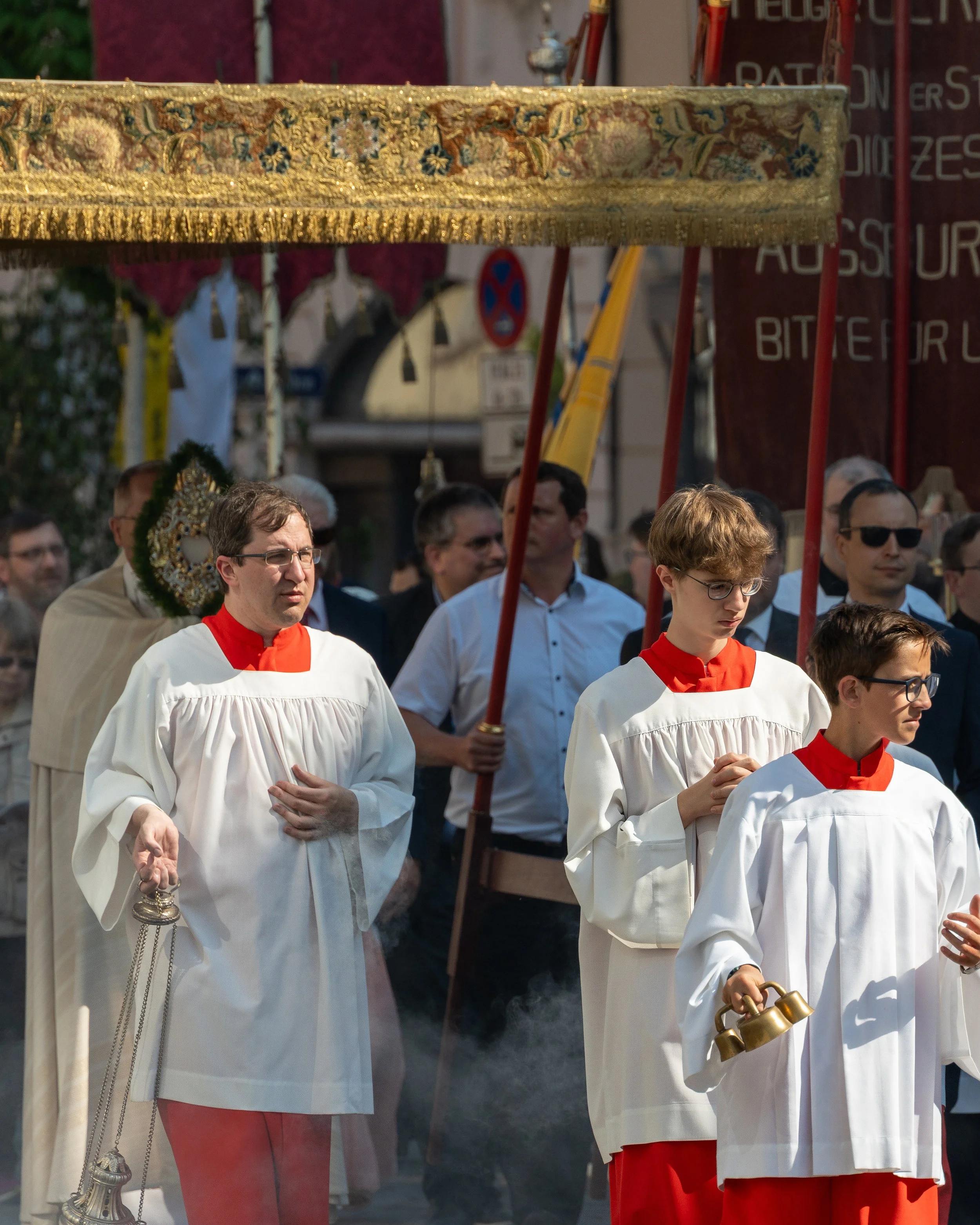 People participating in a religious procession or ceremony, dressed in white robes with red accents, carrying religious items and under a decorated canopy, with onlookers in the background.