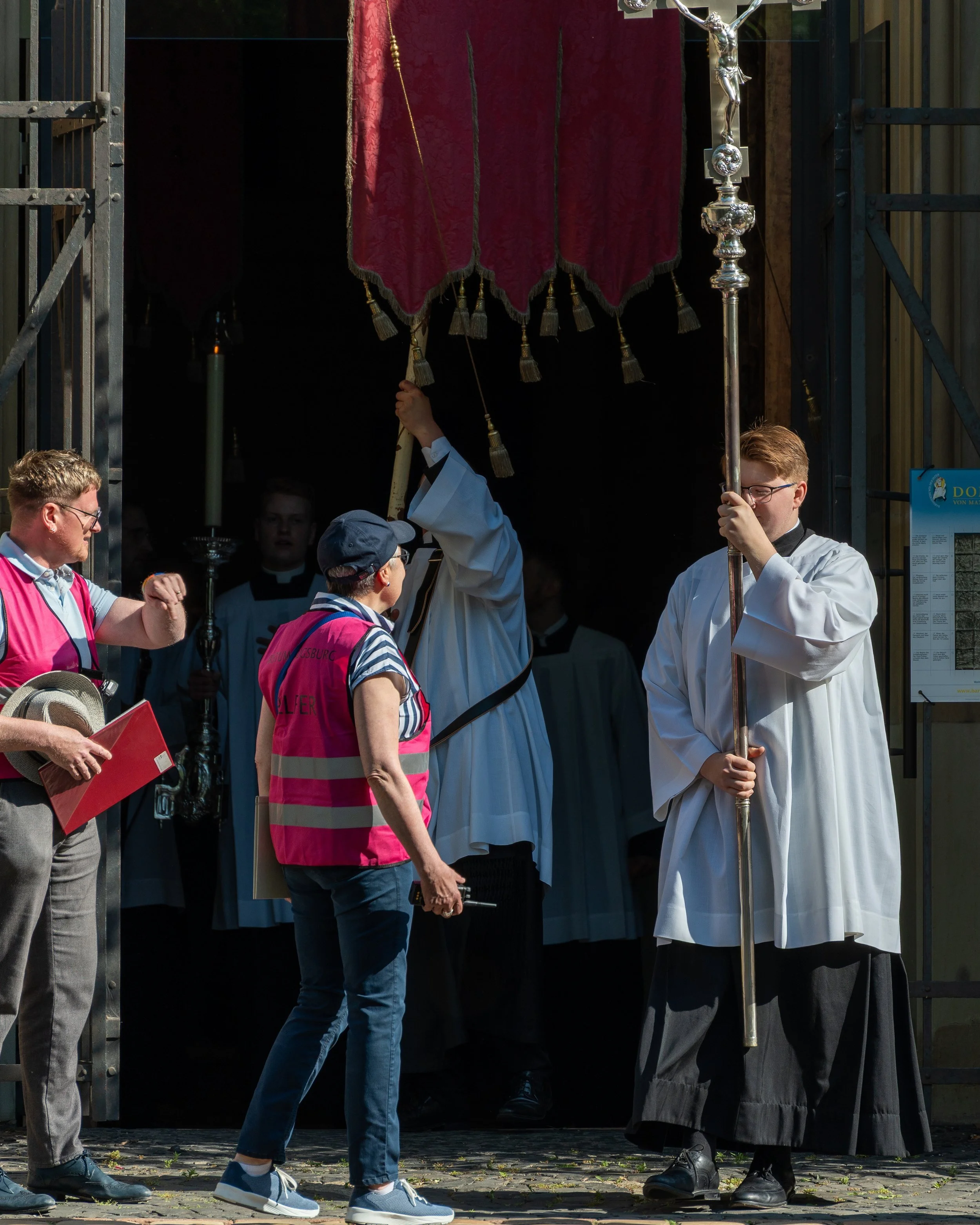 People participating in a religious procession or ceremony, with clergy in white robes holding a cross and a staff, entering a church or chapel through a metal gate. Two women and a man are talking outside, one woman wearing a pink vest and a cap, th