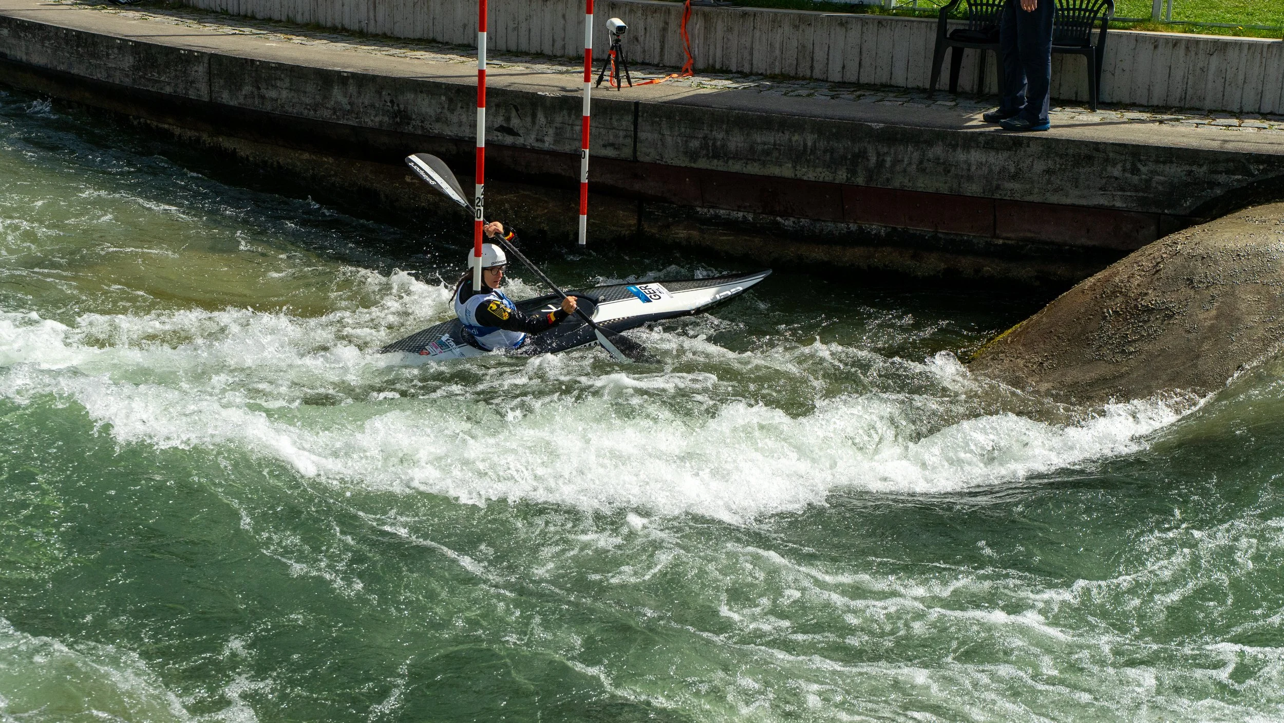 A person in a kayak wearing a helmet navigates through white water rapids near a stone embankment, with a person standing on the embankment observing.