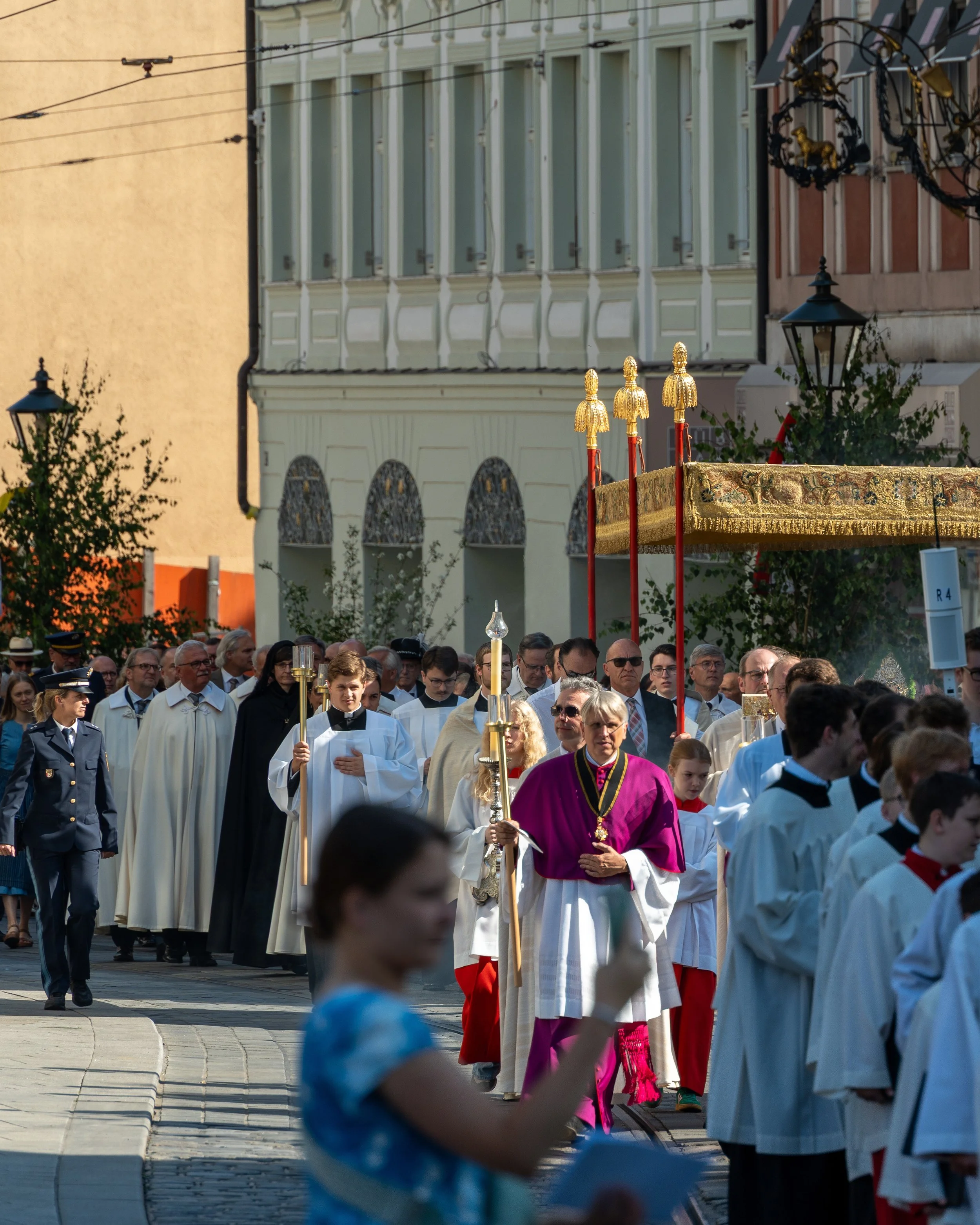 A religious procession with clergy and attendees walking on a street with buildings in the background.