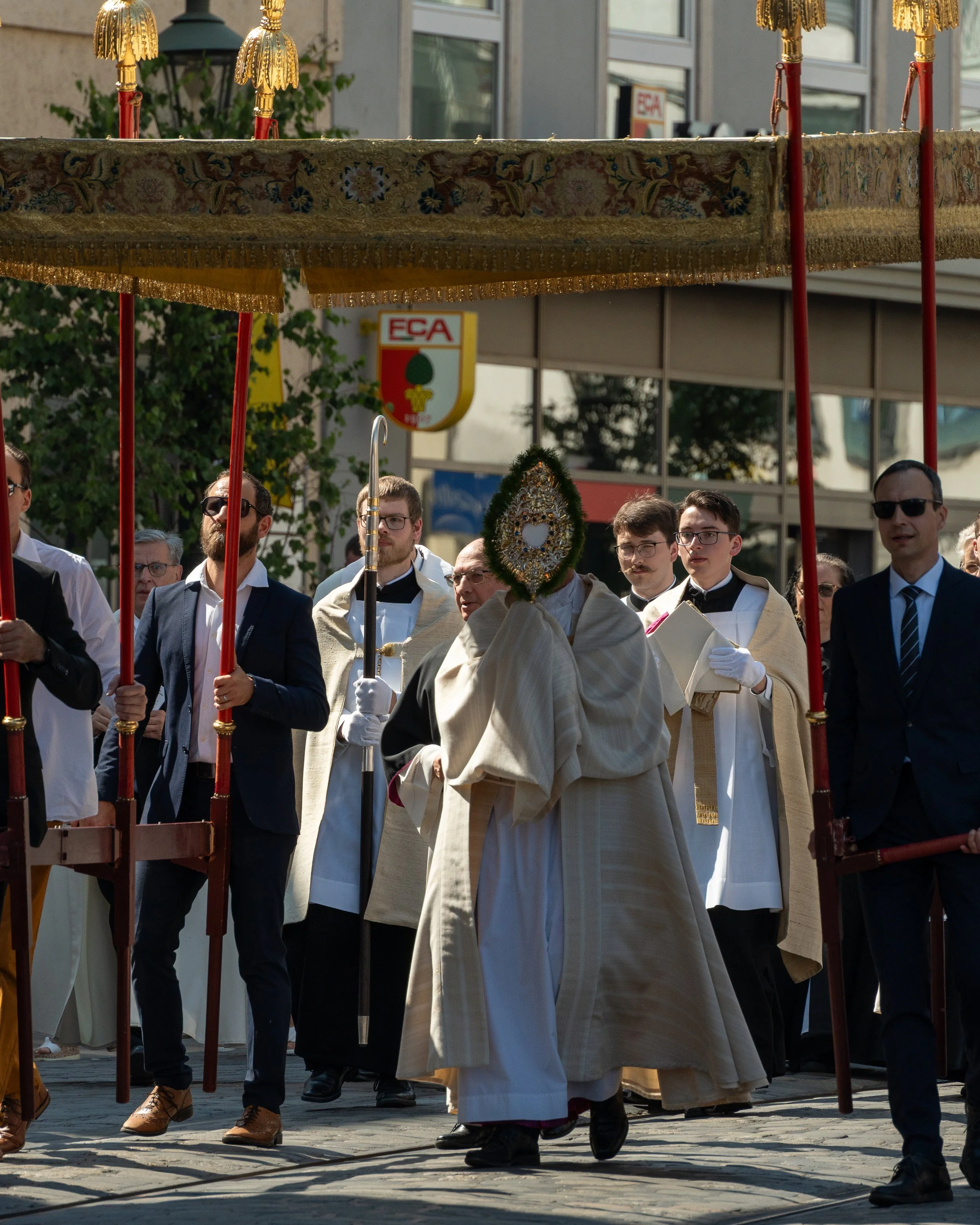 A religious procession with clergy and laypeople, centered around a priest holding a decorated monstrance, under a canopy carried by several men, in an urban setting.