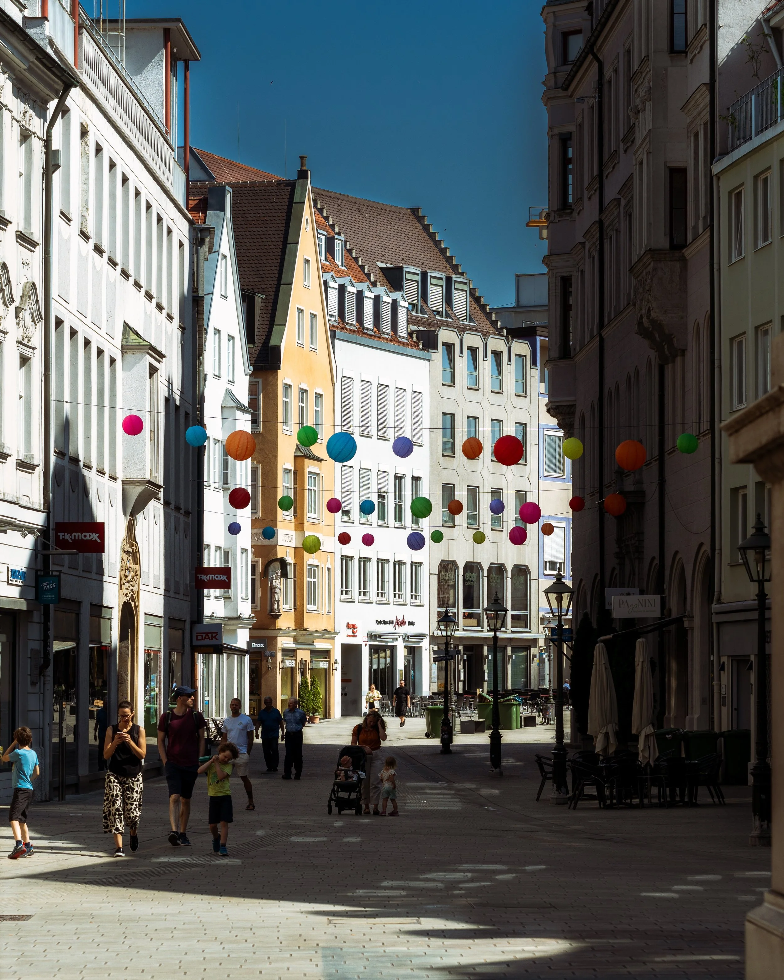 People walking down a colorful city street decorated with hanging paper lanterns under a clear blue sky.