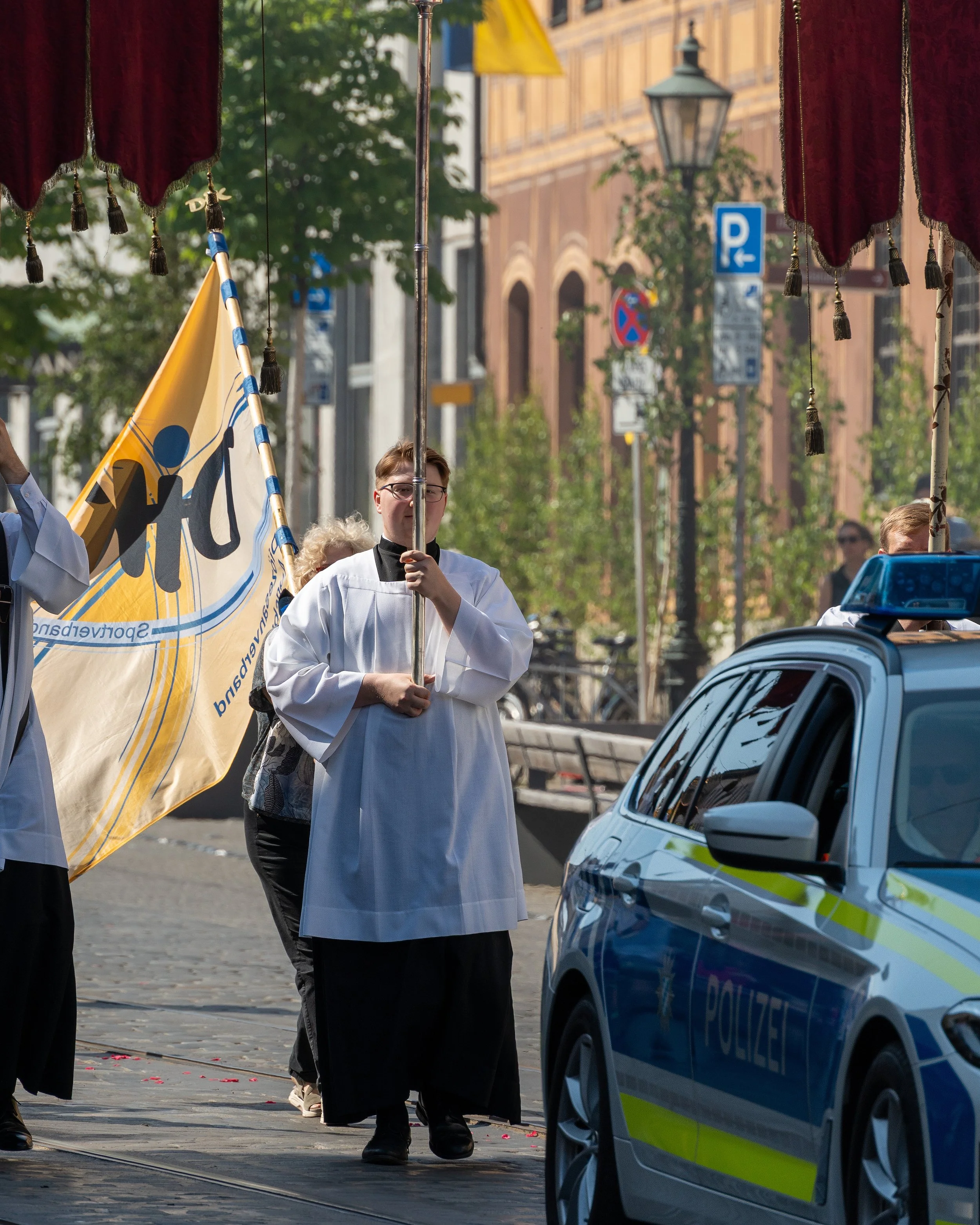 A priest in religious attire holding a yellow flag with black figures and text, standing on a city street next to a police car, during a parade or procession.